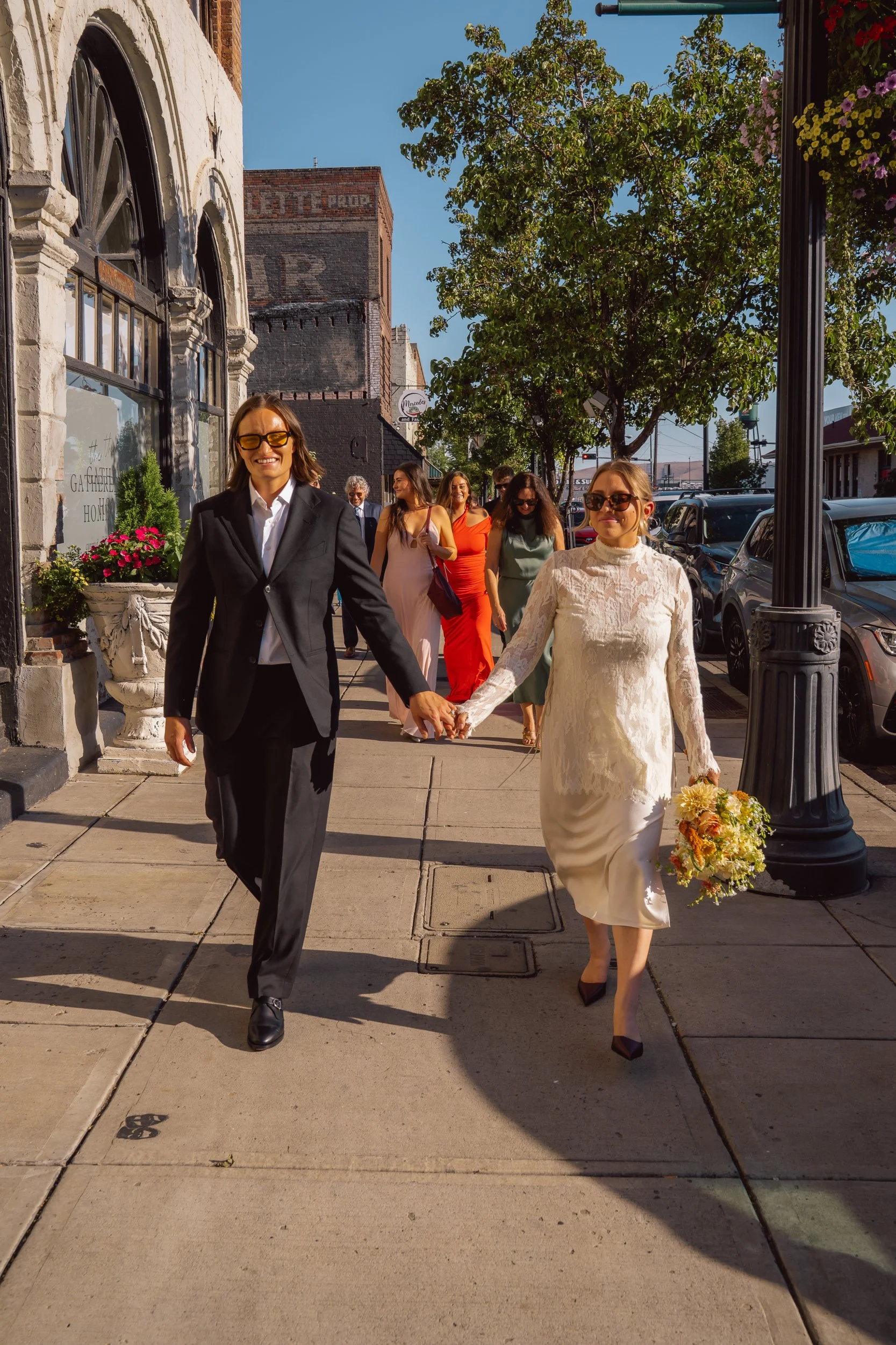 A group of women walking on a city sidewalk, with the woman in the front wearing a white lace dress and holding a bouquet, while another woman next to her wears a black suit. Several other women in colorful dresses follow behind them, and they are sm