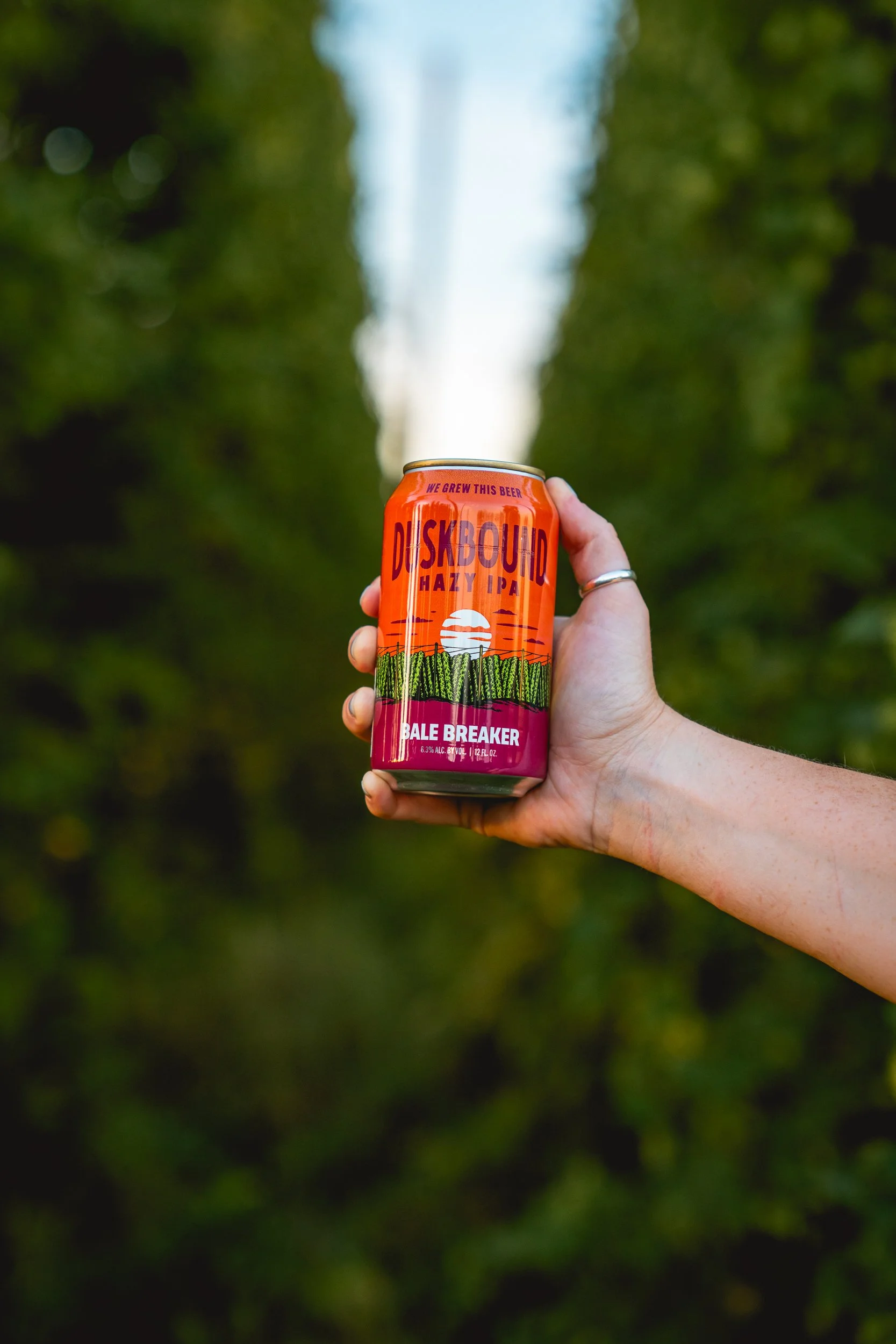 Hand holding an orange can of Duskyound Hazy IPA beer outdoors with green trees in the background.