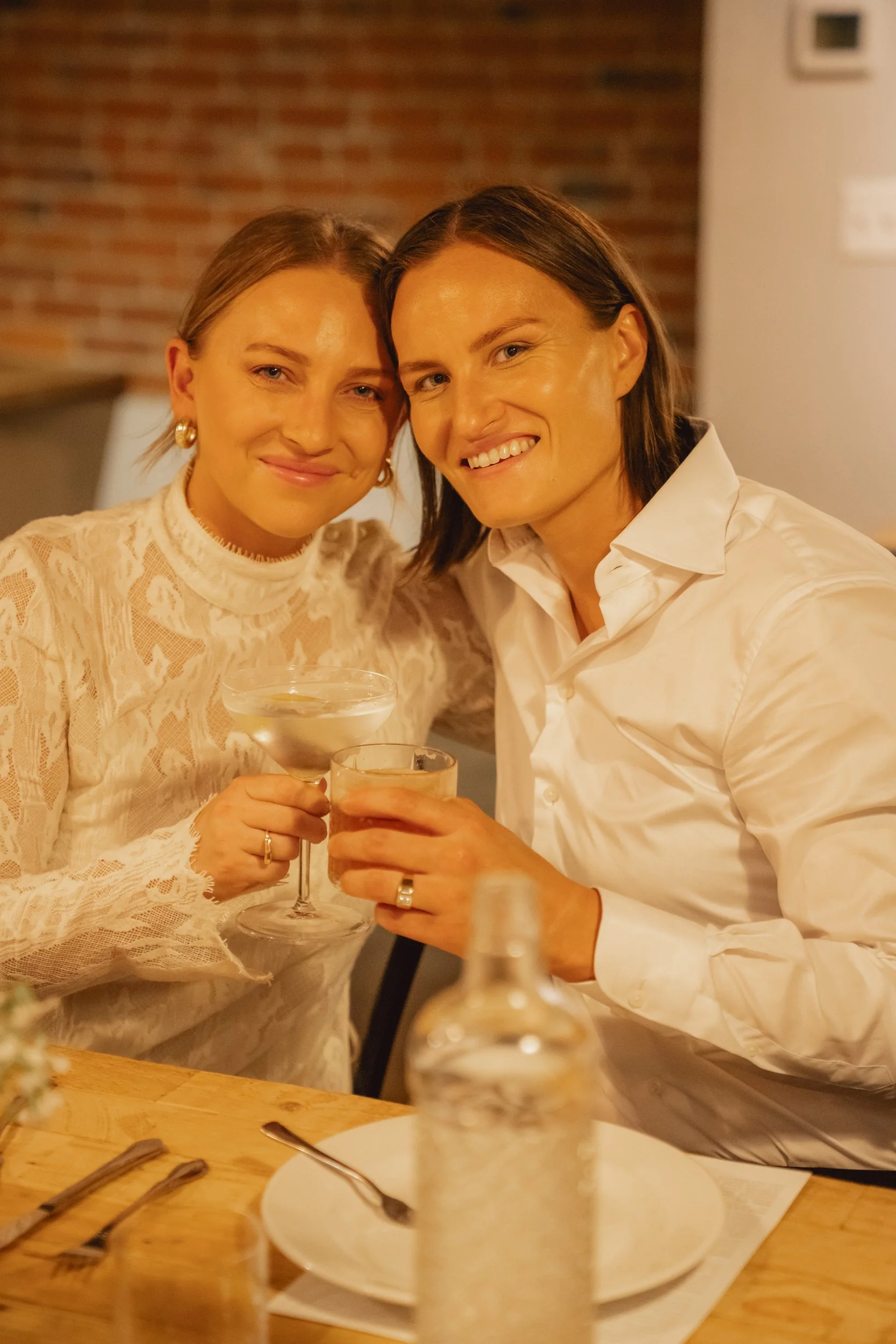 Two women smiling and toasting drinks in a cozy restaurant with a brick wall background.