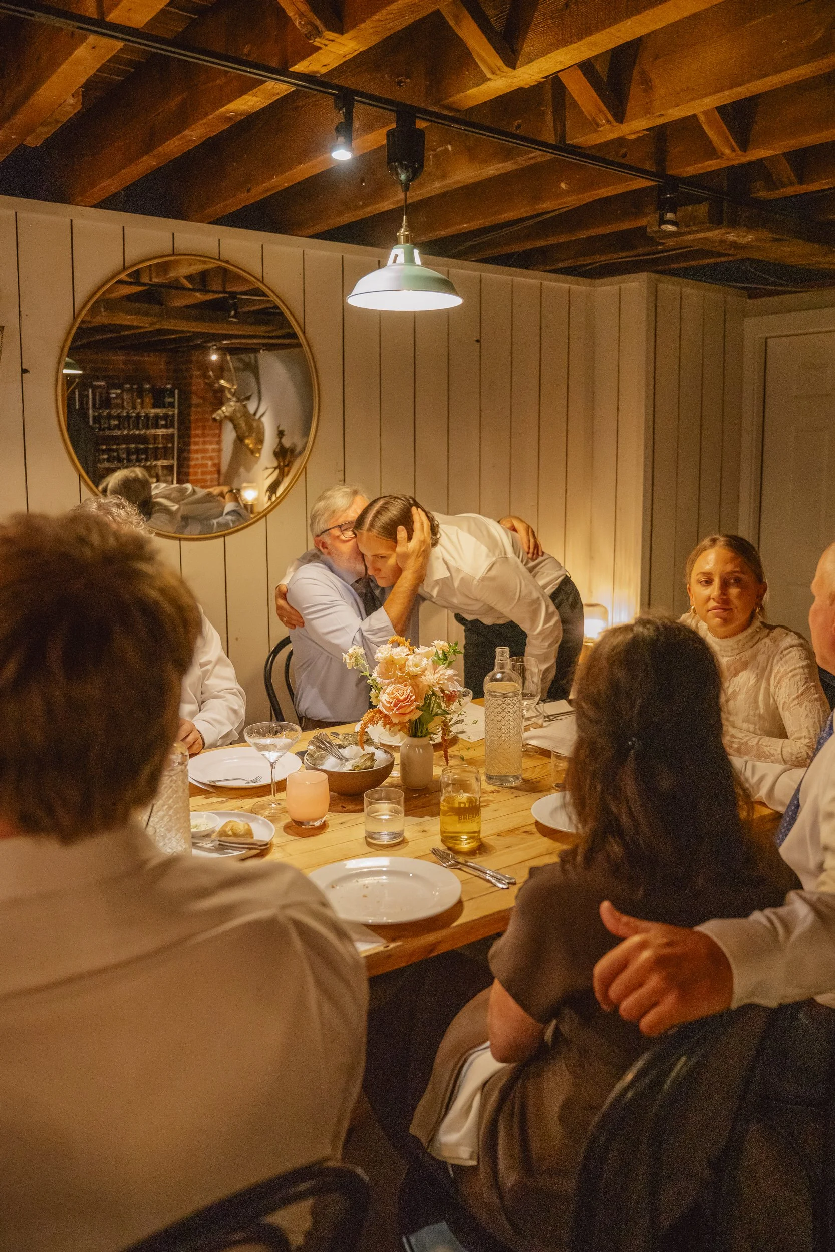 People sitting around a wooden dining table in a cozy, rustic restaurant. Two individuals are embracing, one kissing the other's head. The table is set with plates, glasses, and a floral centerpiece. A mirror on the wall reflects a mounted deer head 