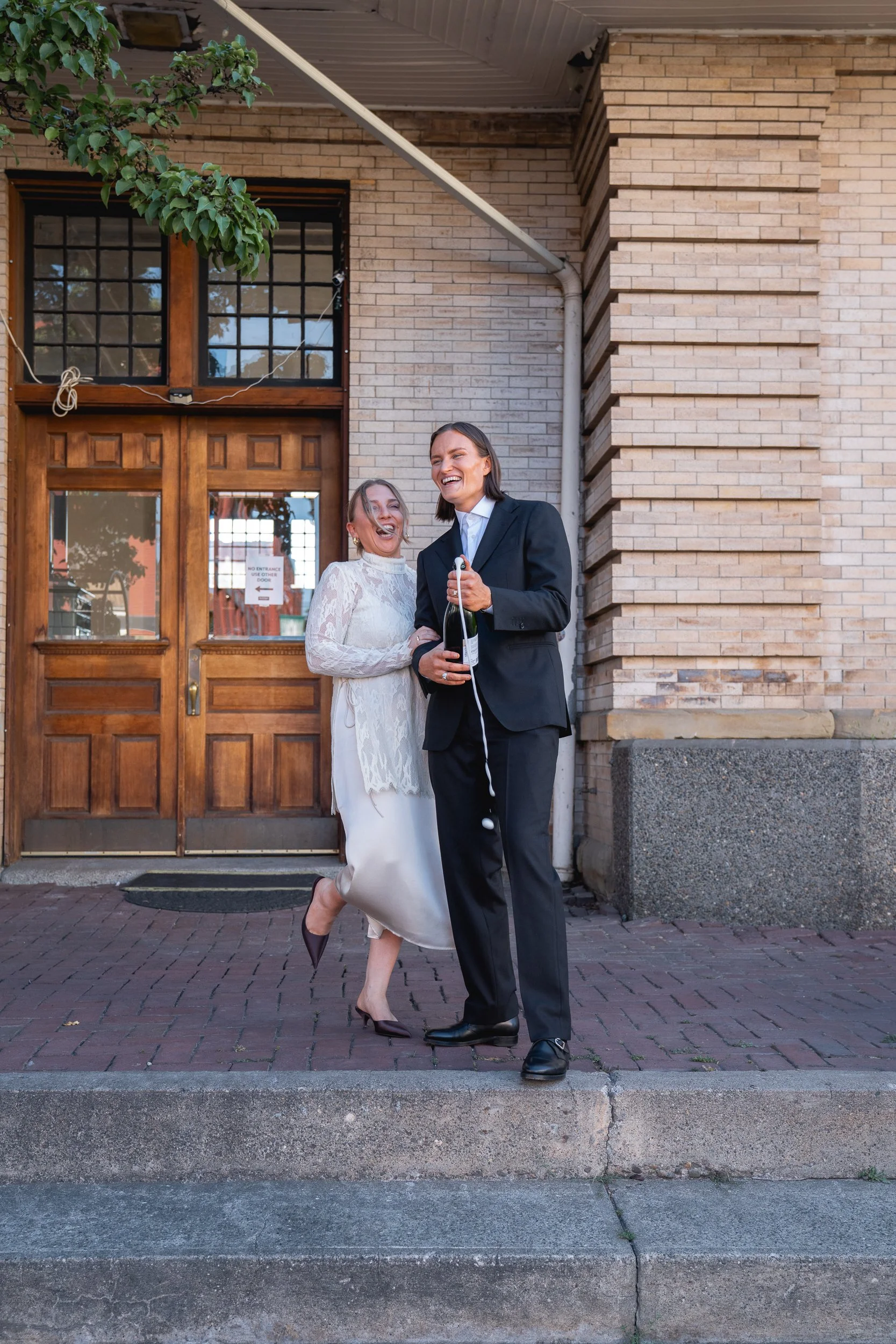 Two women, one in a wedding dress and the other in a black suit, celebrating outside a brick building with wooden doors. They are smiling and holding a bottle of champagne.