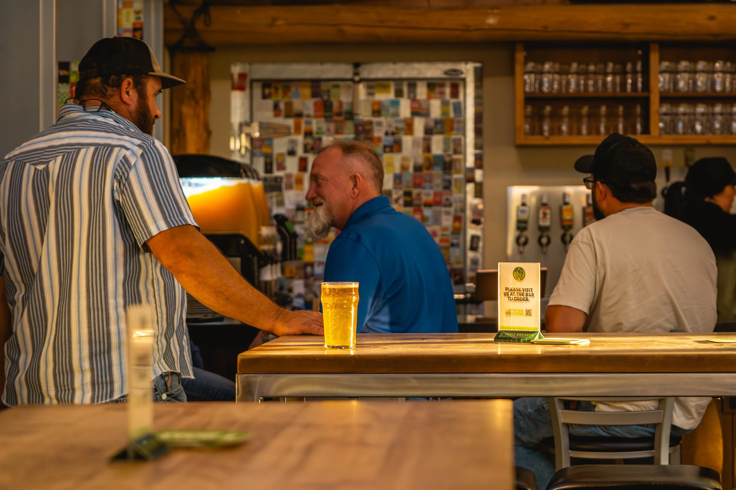 A man in a striped shirt and cap ordering a drink at a bar counter, with a glass of beer in front of him, inside a cozy bar or cafe with warm lighting and wooden decor, and other patrons sitting at the bar.