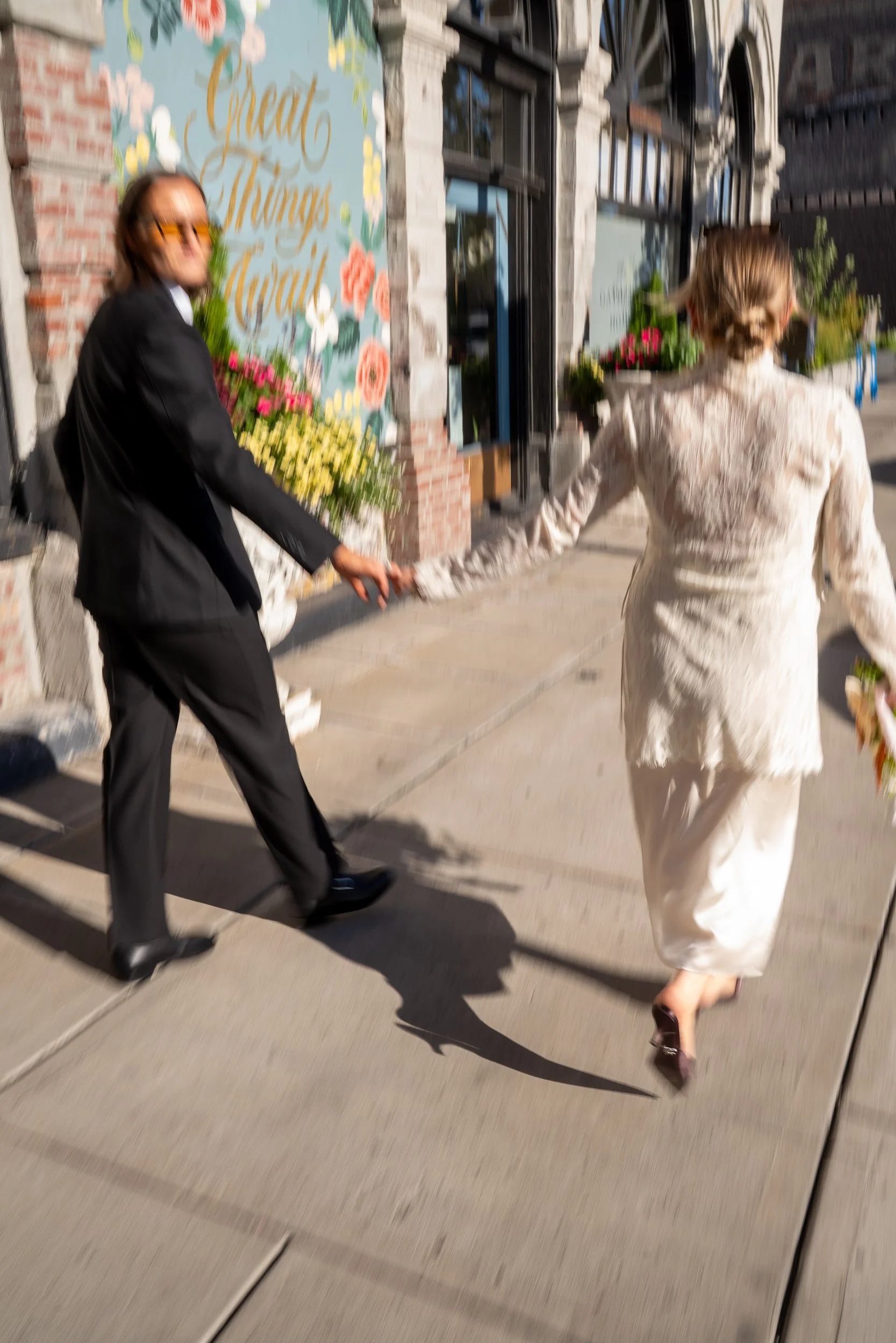 Two women holding hands, walking on sidewalk outside a floral shop with a sign that reads "Great Things Ahead." One woman is dressed in a black suit and sunglasses, while the other wears a cream lace dress and heels.