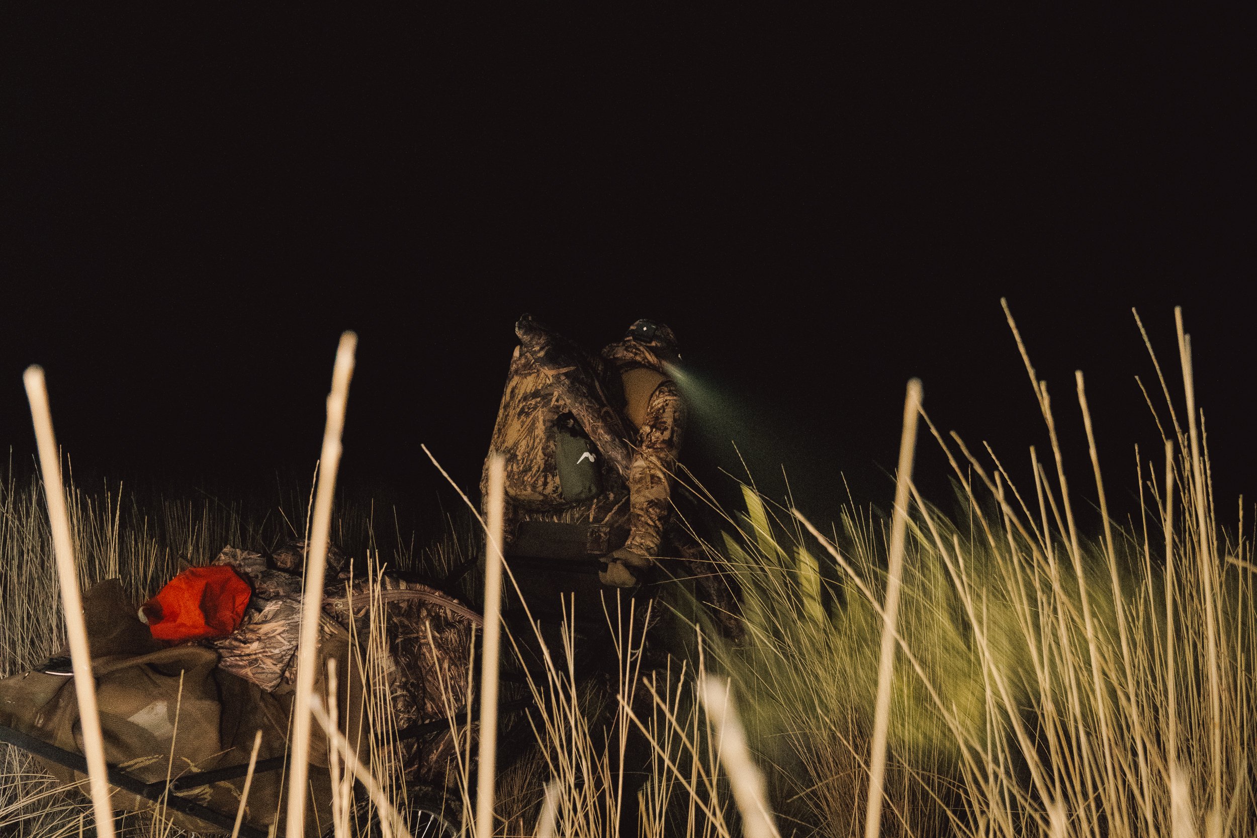 A person in camouflage clothing riding a bicycle with a large backpack, illuminated by a headlamp, through tall grass at night.