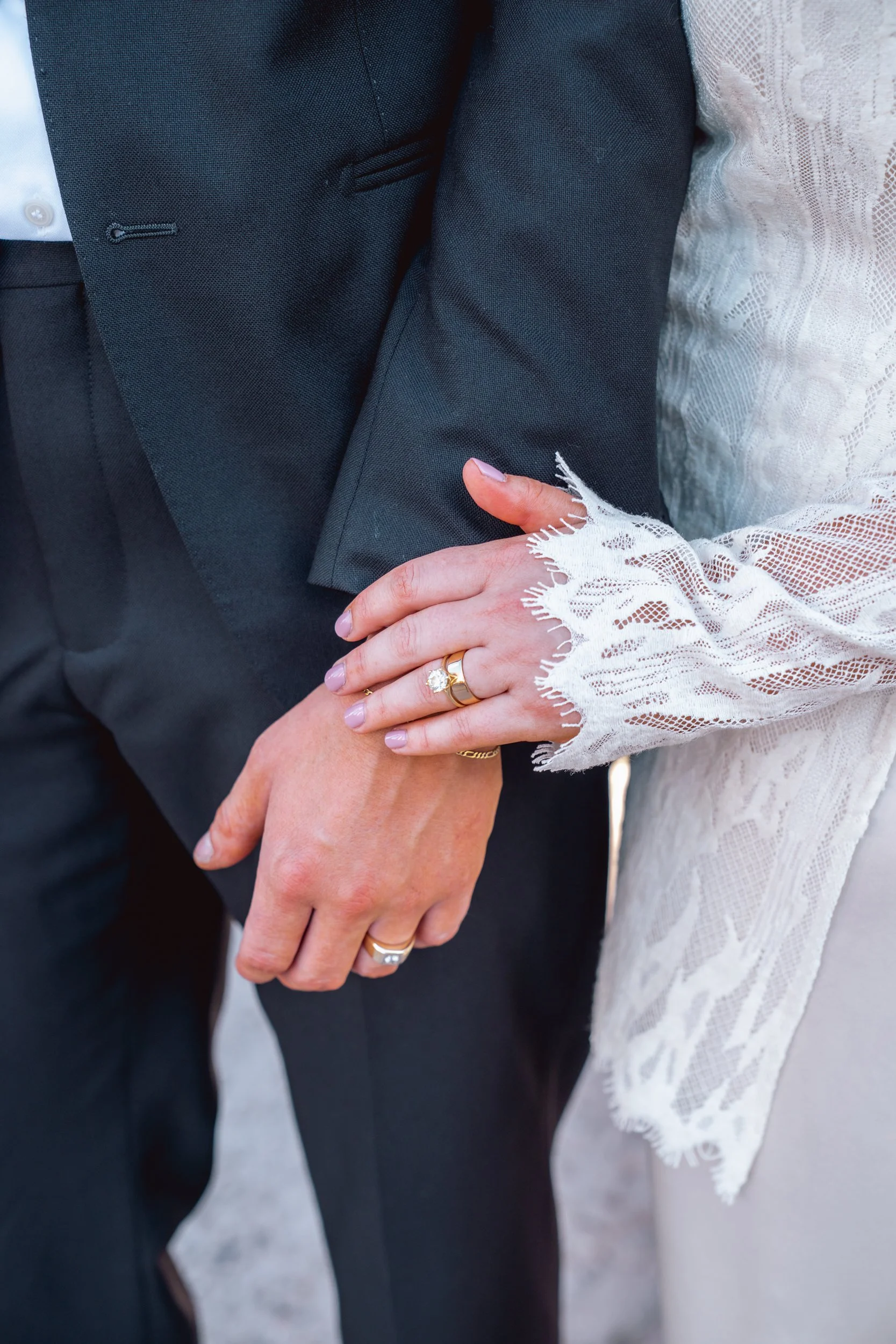 Close-up of a bride and groom holding hands, with the bride wearing lace sleeves and wedding rings, and the groom in a black suit.