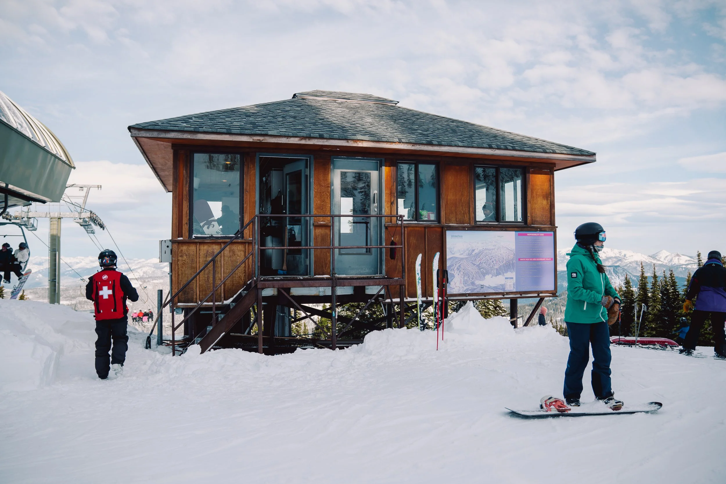 Ski resort scene with people skiing on snow, a wooden building with large windows, and a snow-covered mountain landscape in the background.