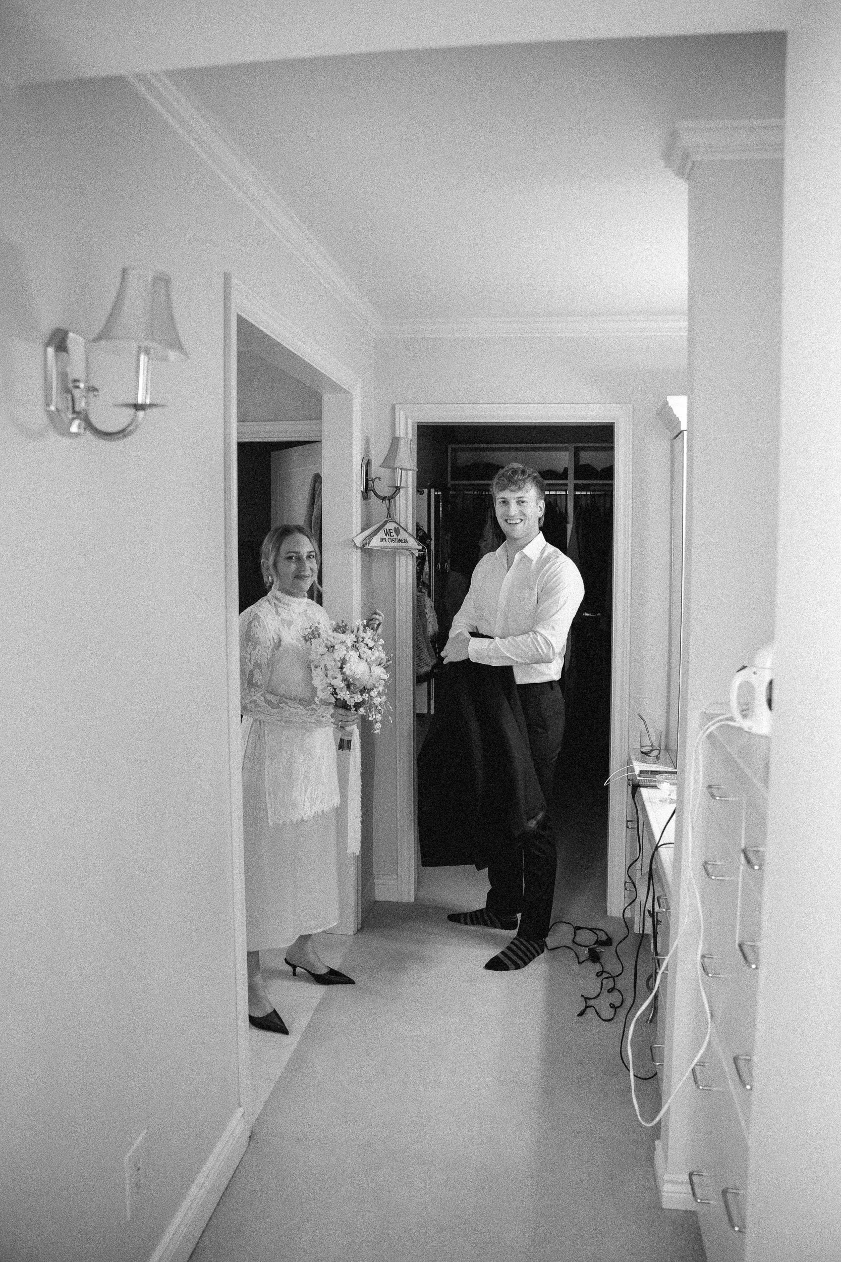 A woman in a white dress holding a bouquet of flowers and a man in a white shirt, both smiling, standing in a hallway with open closet in the background.