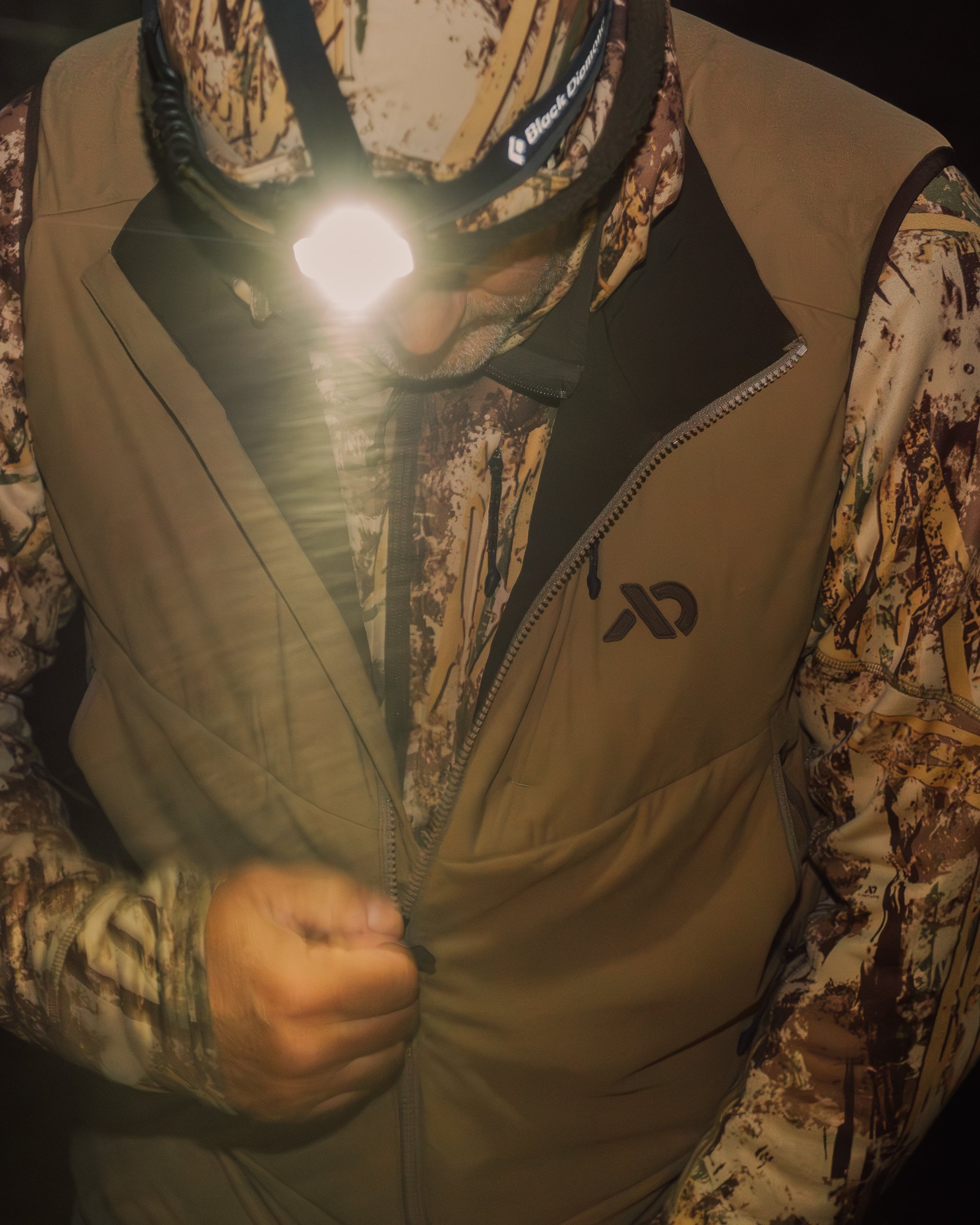 A person wearing camouflage clothing and a tan vest, with a headlamp turned on.
