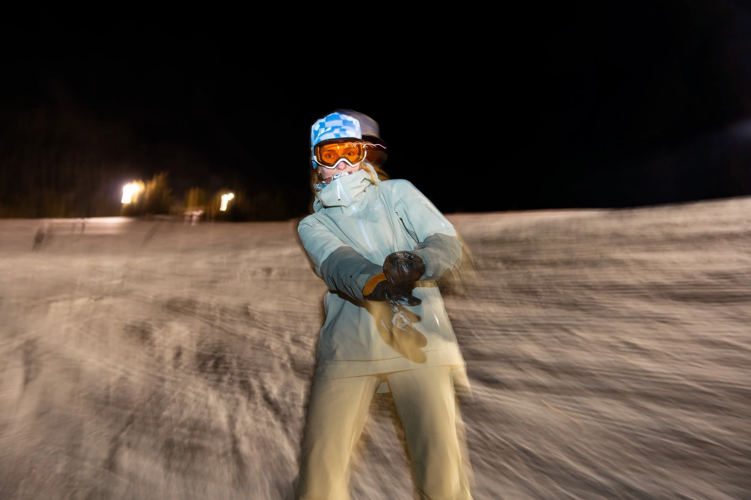 A person skiing at night wearing a beige jacket, beige ski pants, goggles, a helmet, and gloves on a snow-covered slope.