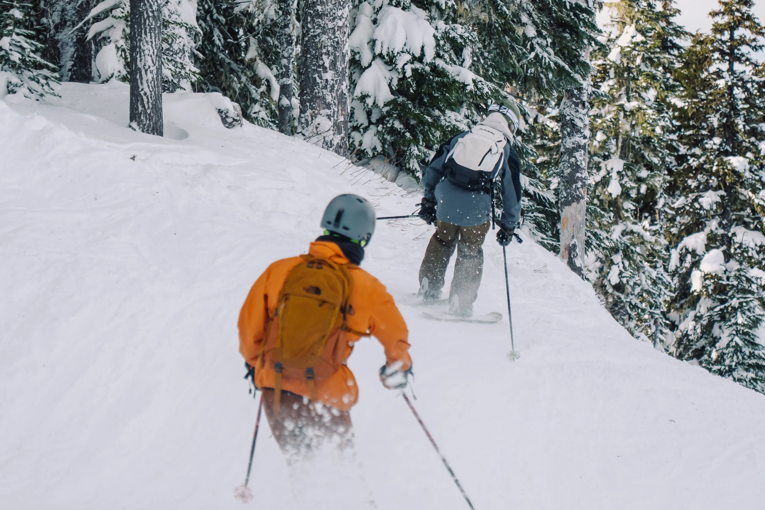 Two people snowboarding through a snowy forest path surrounded by snow-covered evergreen trees.