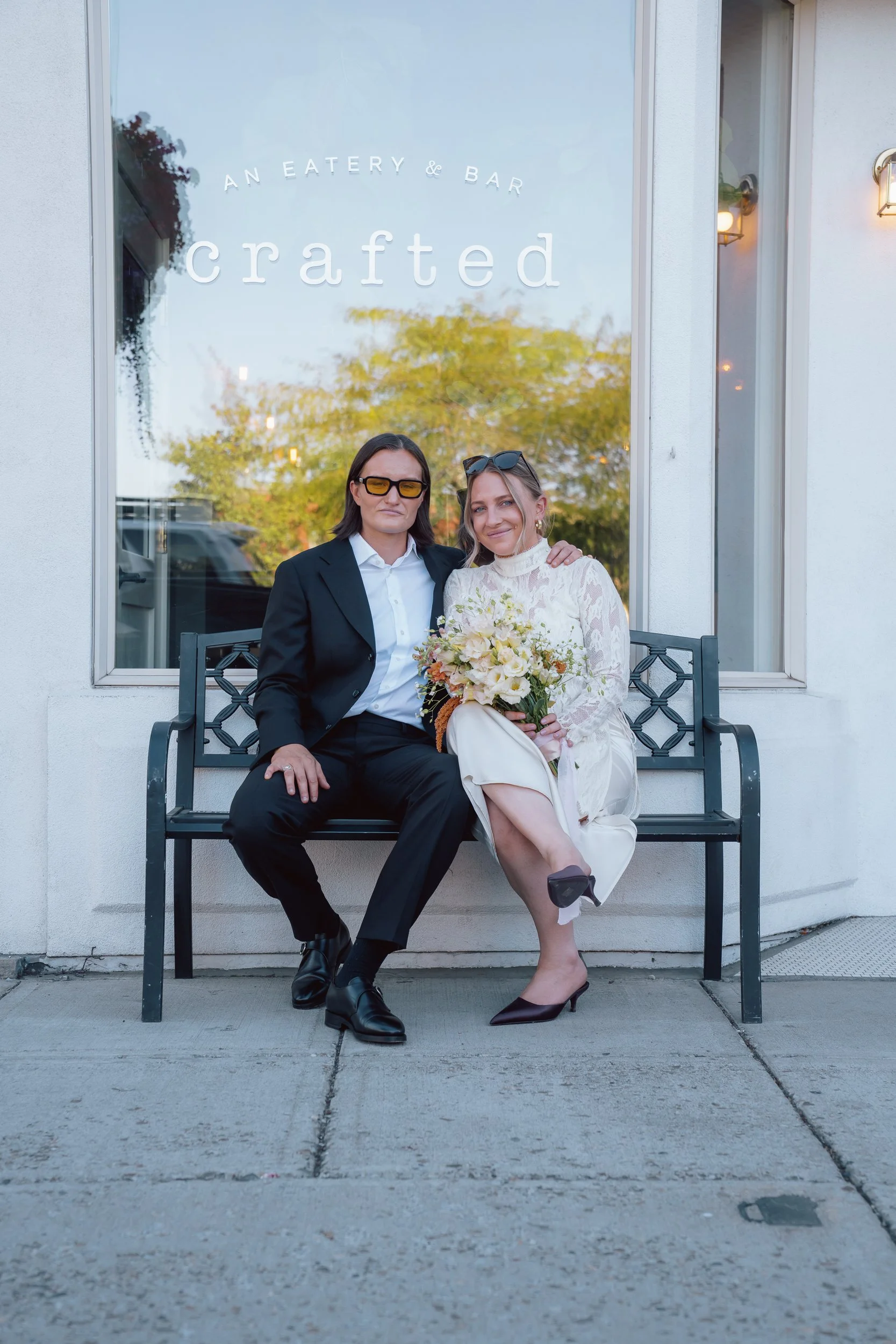 Two women sitting on a bench outside a restaurant with a sign that says "An Eatery & Bar" and "crafted." One woman is wearing a black suit and yellow glasses, and the other woman is wearing a white dress, holding a bouquet of flowers, and has sunglas