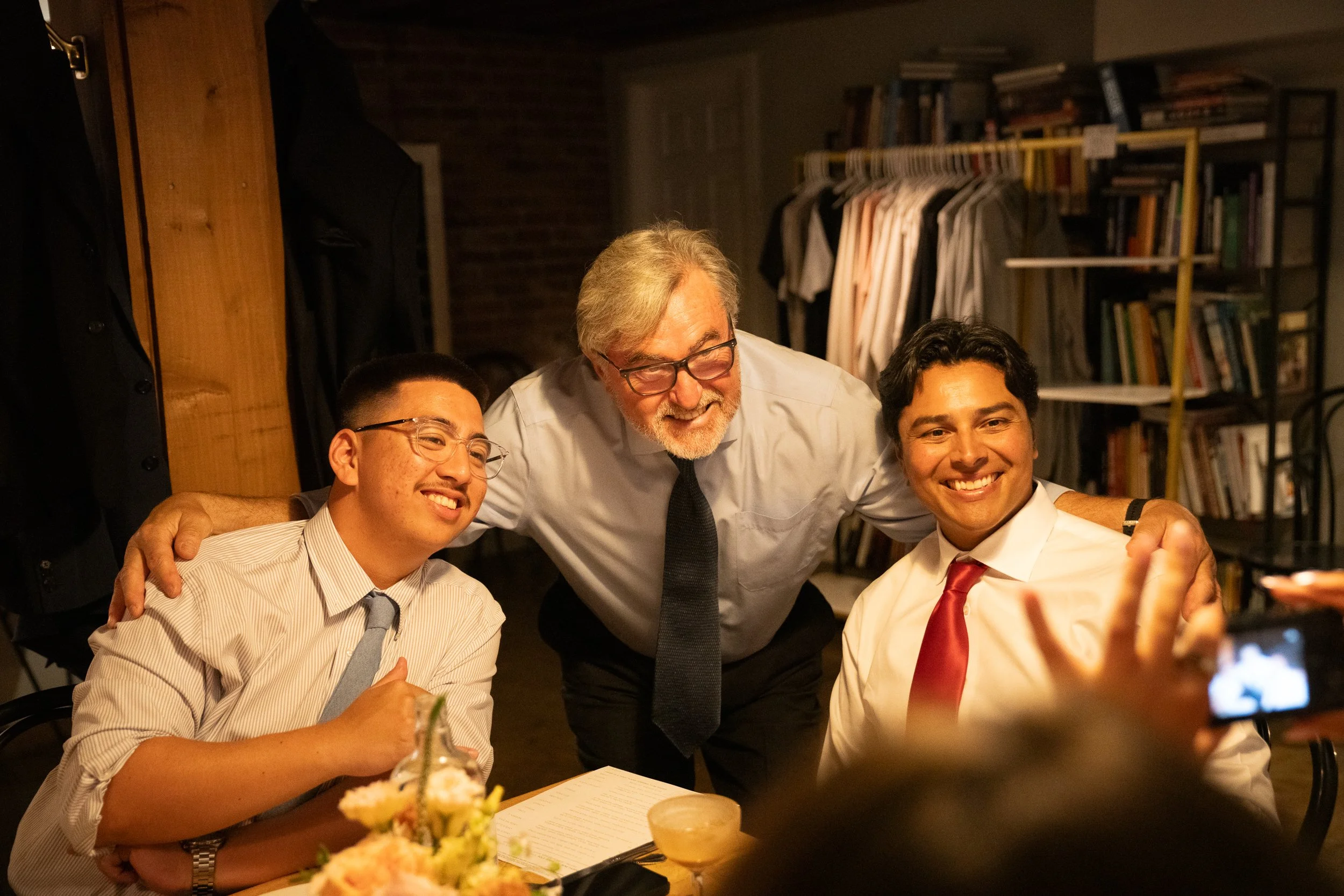 Three men, two seated and one standing, smiling and taking a photo together in a room with bookshelves and clothing racks in the background.
