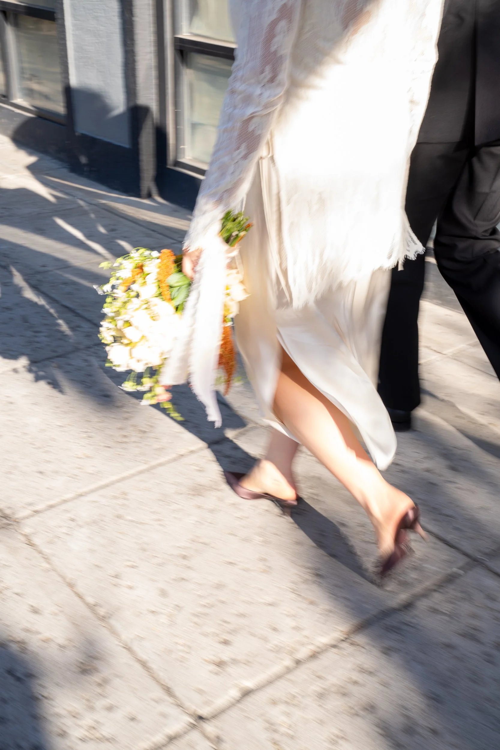 A bride walking on a sidewalk holding a bouquet of flowers, wearing a white dress and heels.