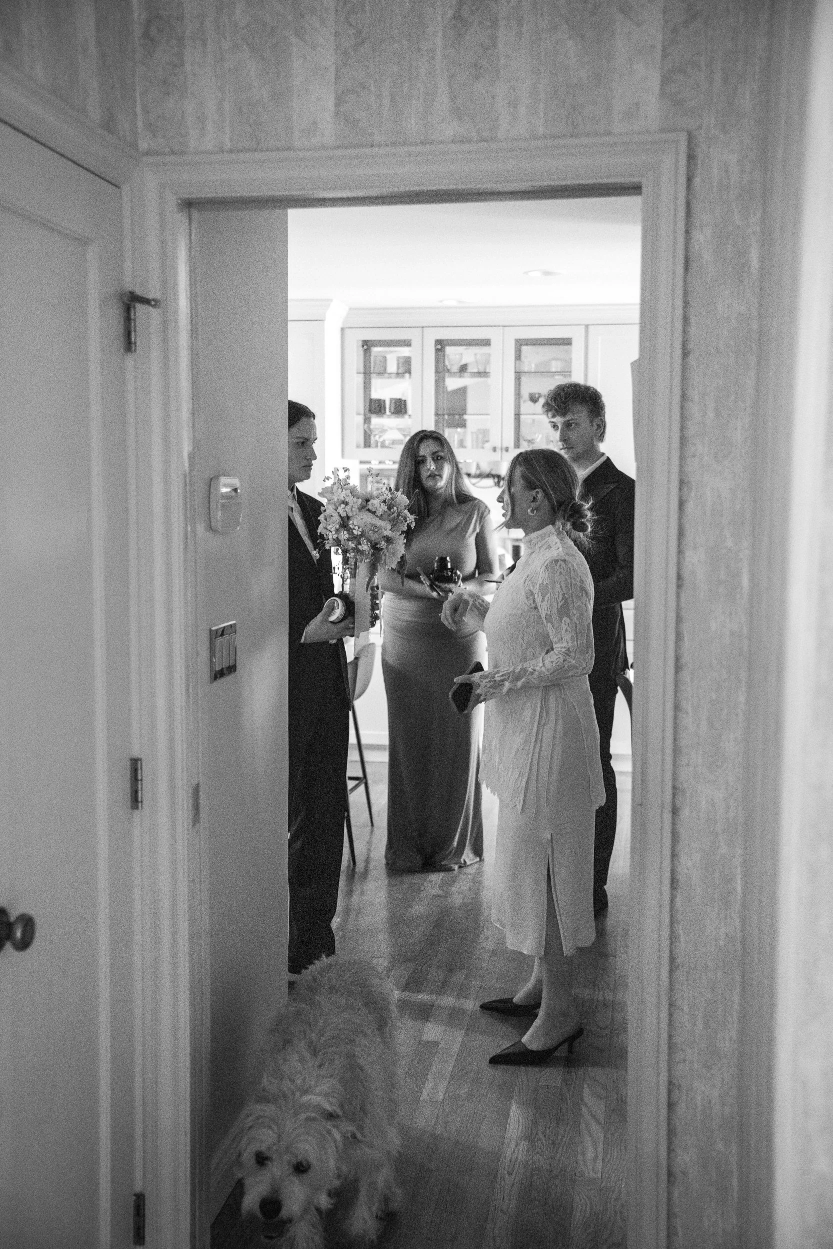 A black and white photo of four people standing in a room, having a conversation. Two women and two men are visible, one woman is holding a bouquet of flowers. A dog is in the foreground, near the doorway.