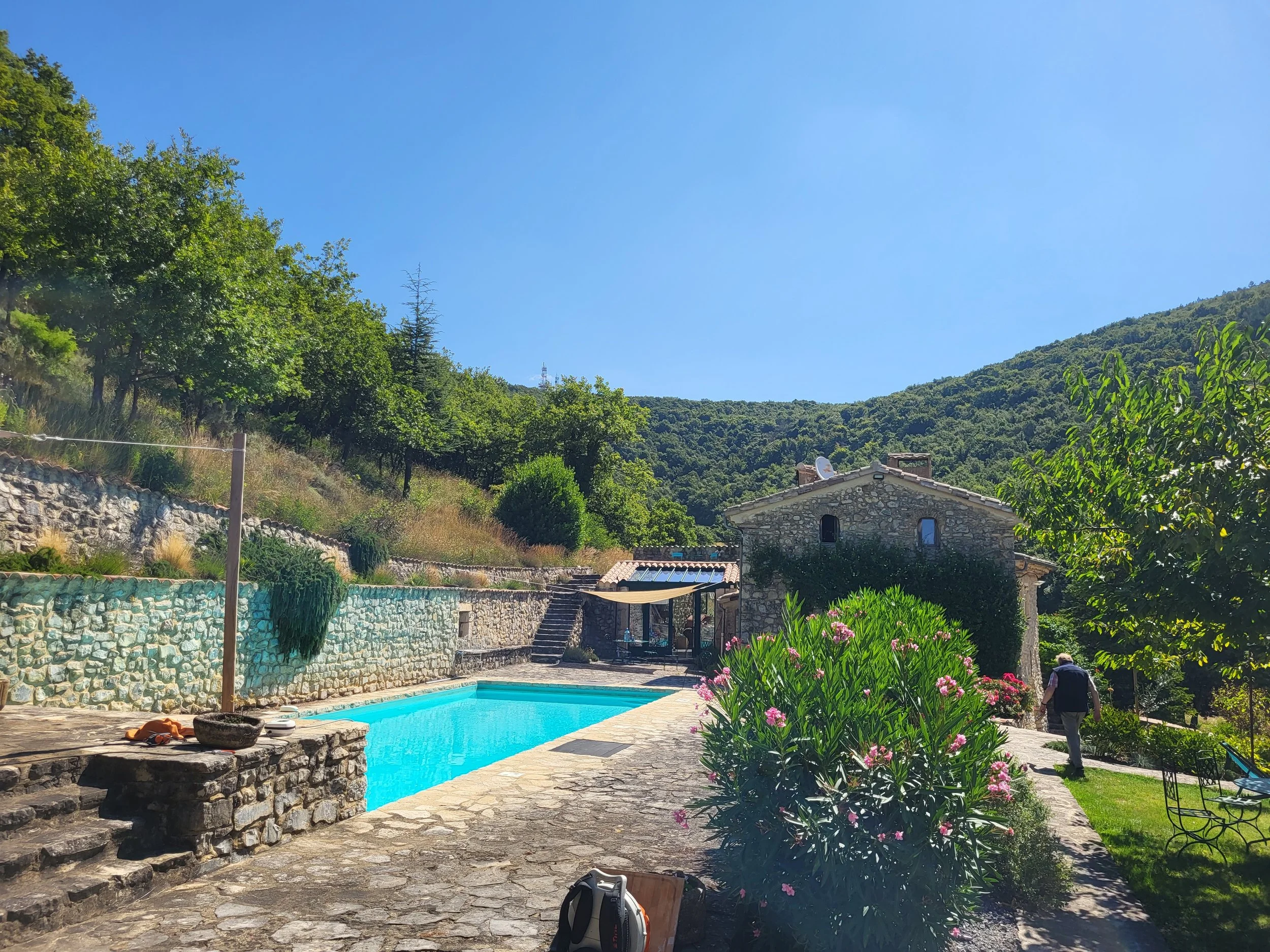 A sunny outdoor scene featuring a stone house next to a swimming pool, surrounded by greenery and hills under a clear blue sky.