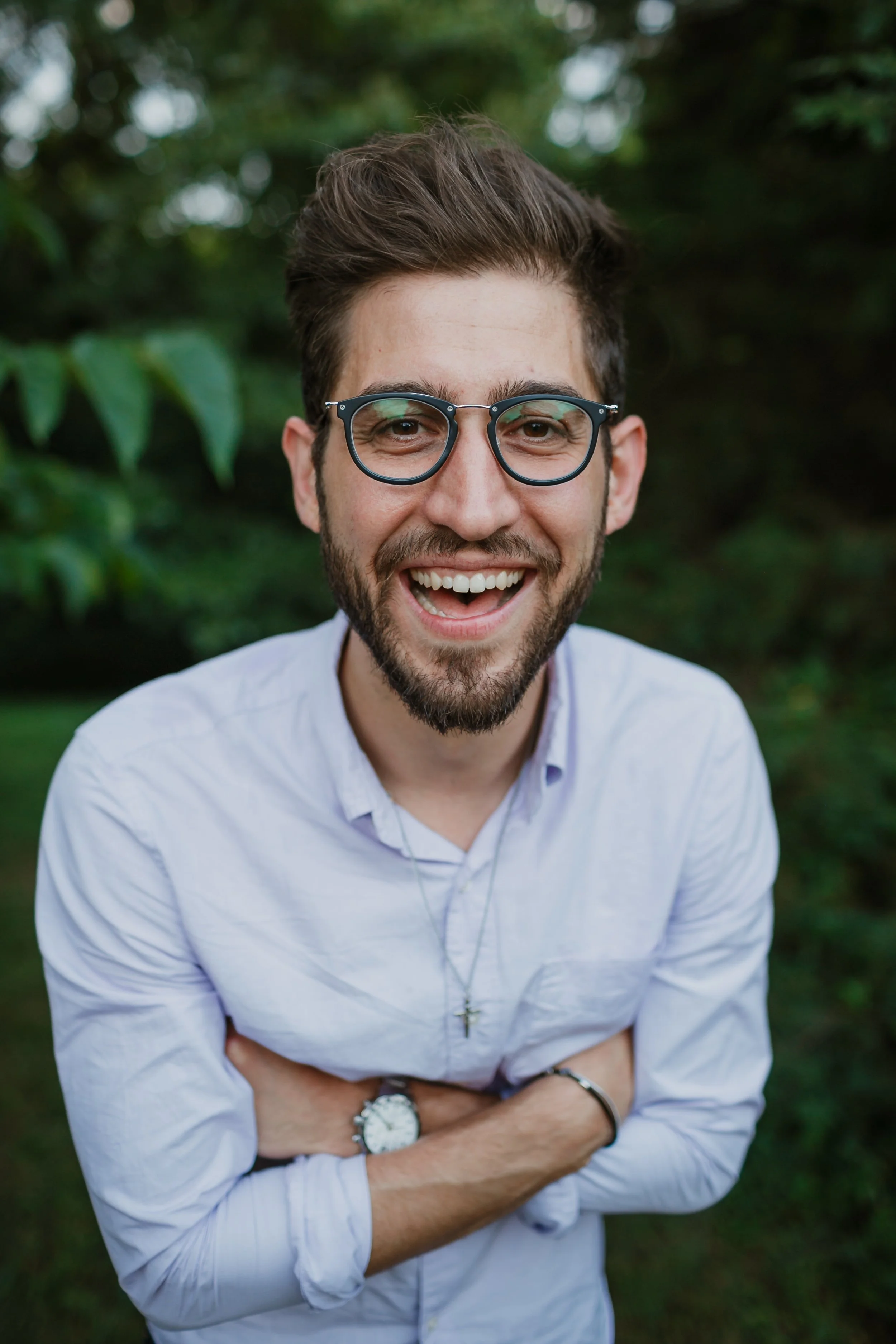 A young man with glasses and a beard smiling outdoors in front of greenery, wearing a white shirt and a watch.