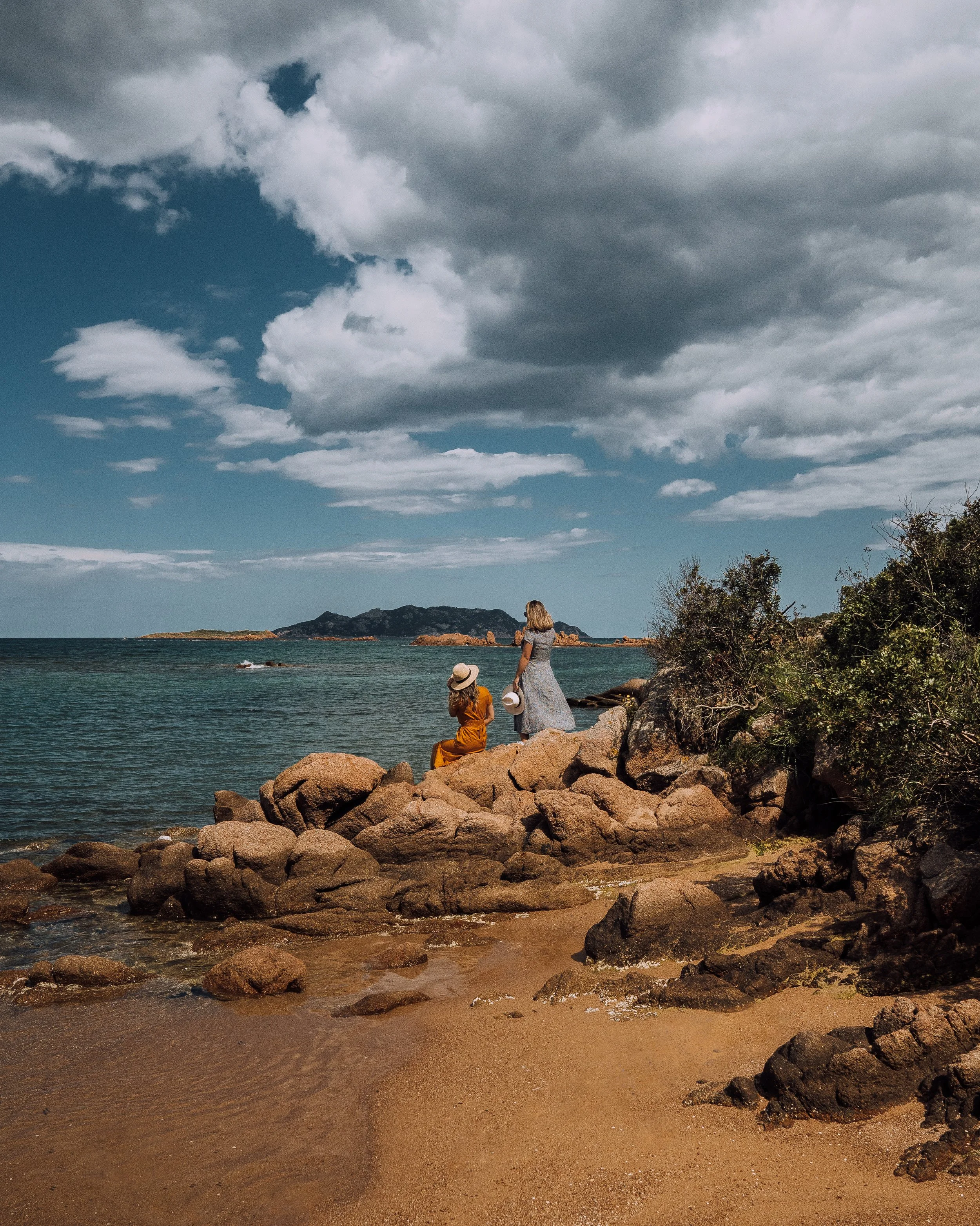 Two women wearing dresses and hats sitting on coastal rocks near the sea with a cloudy sky overhead.