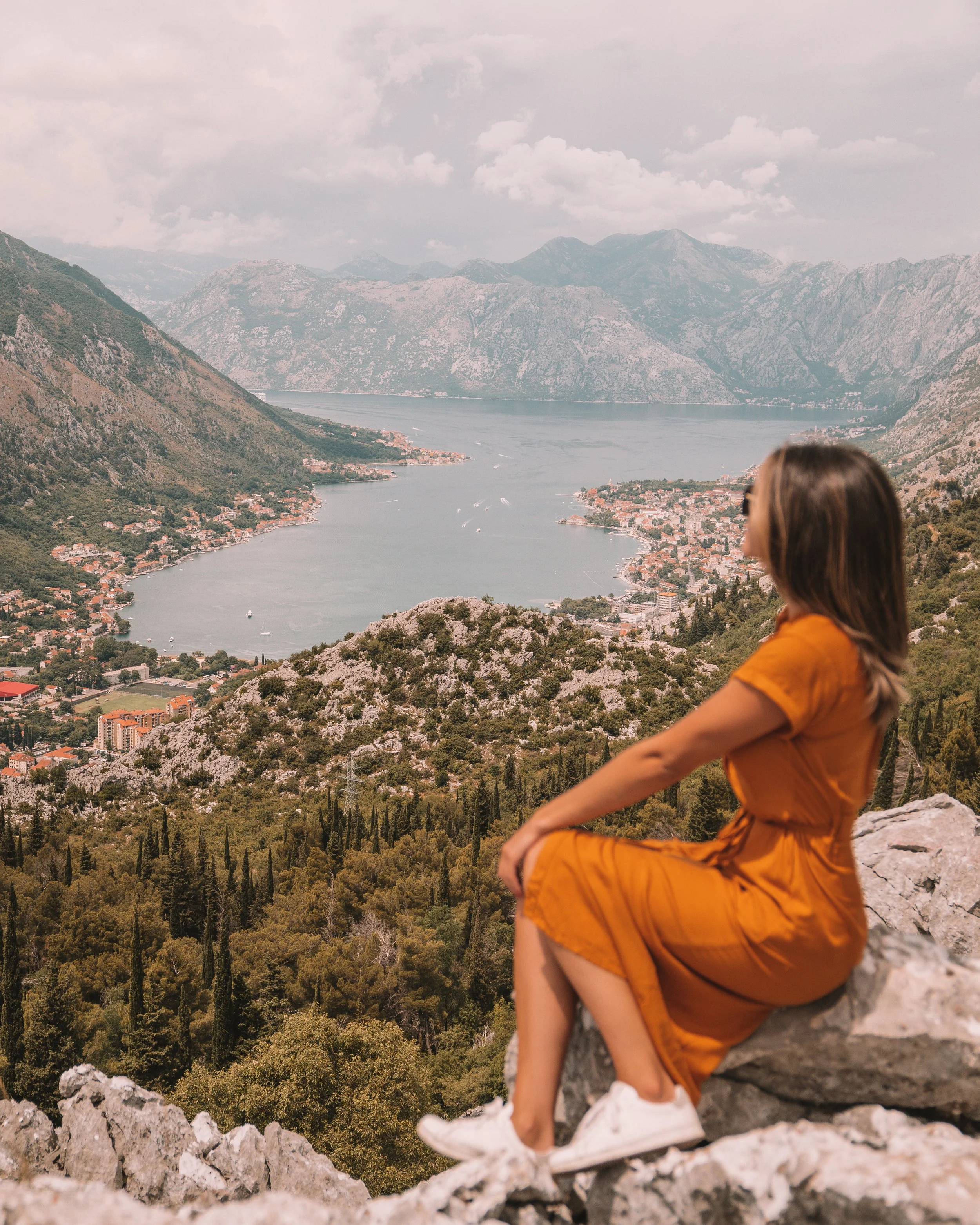 Person in orange dress sitting on rocky terrain, overlooking a scenic view of a bay with mountains and village, under a cloudy sky.