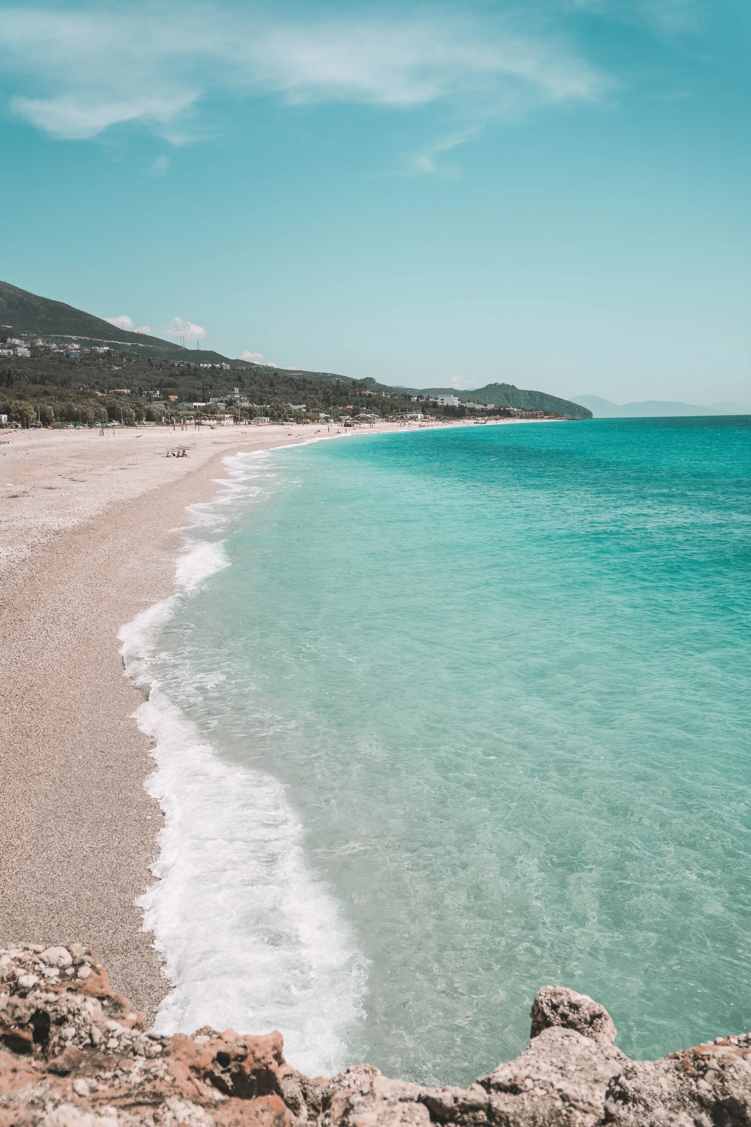 Scenic beach with turquoise water and waves gently hitting a sandy shore, surrounded by lush green hills and a clear blue sky.