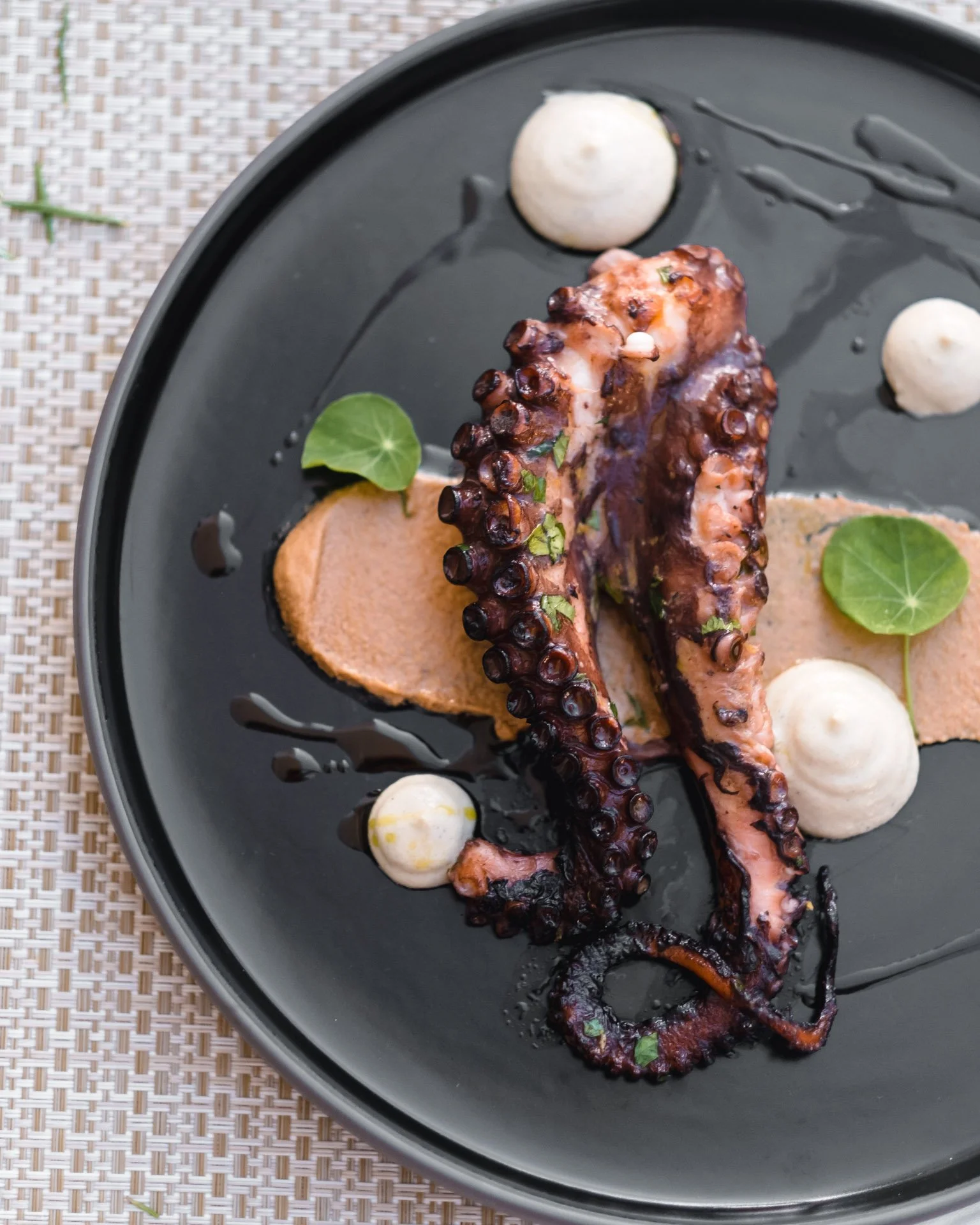 Grilled octopus tentacle served on a black plate with decorative sauces and garnished with green leaves.
