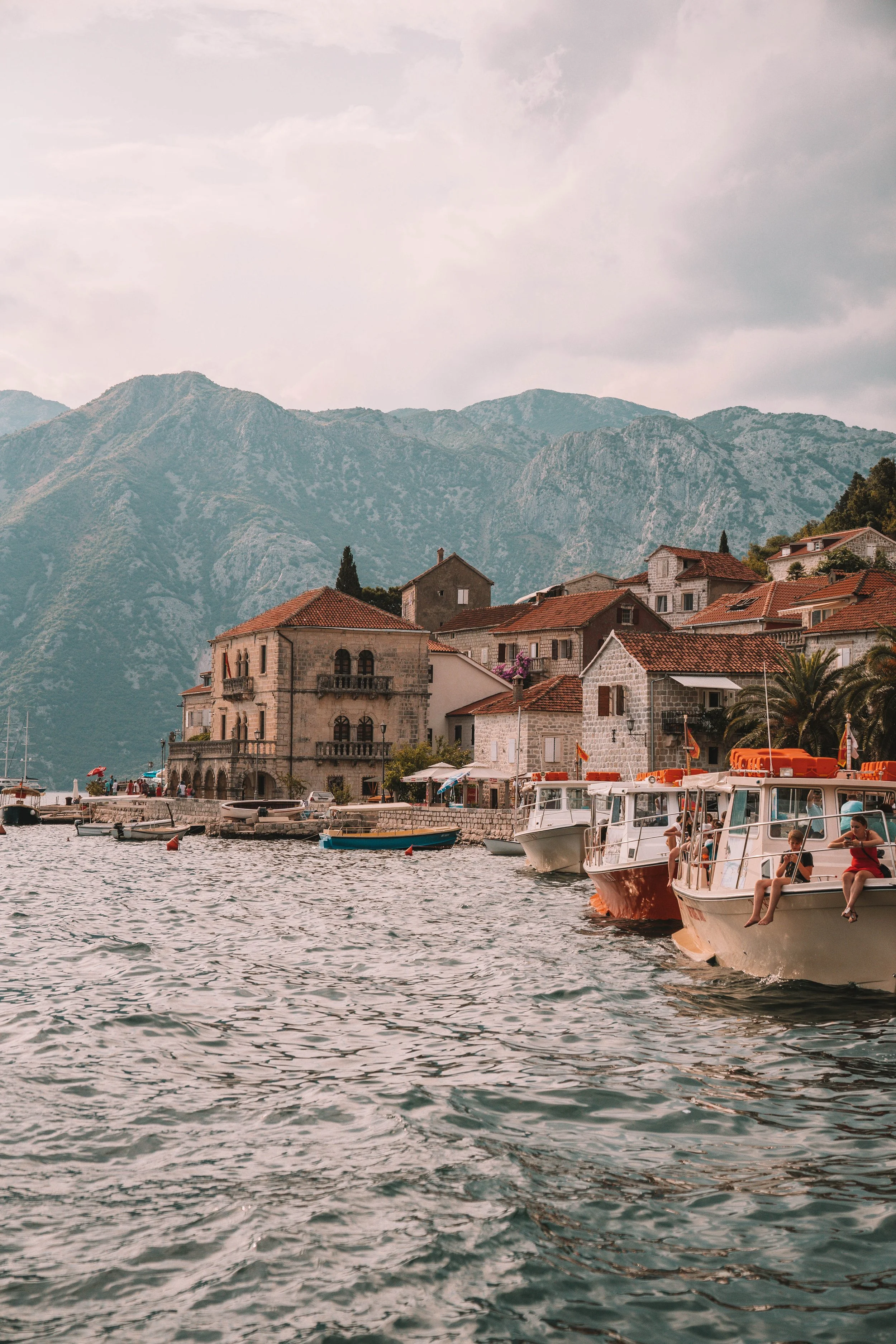 Scenic view of a coastal village with traditional stone buildings, red-tiled roofs, and boats moored in the water. Mountainous backdrop and cloudy sky.