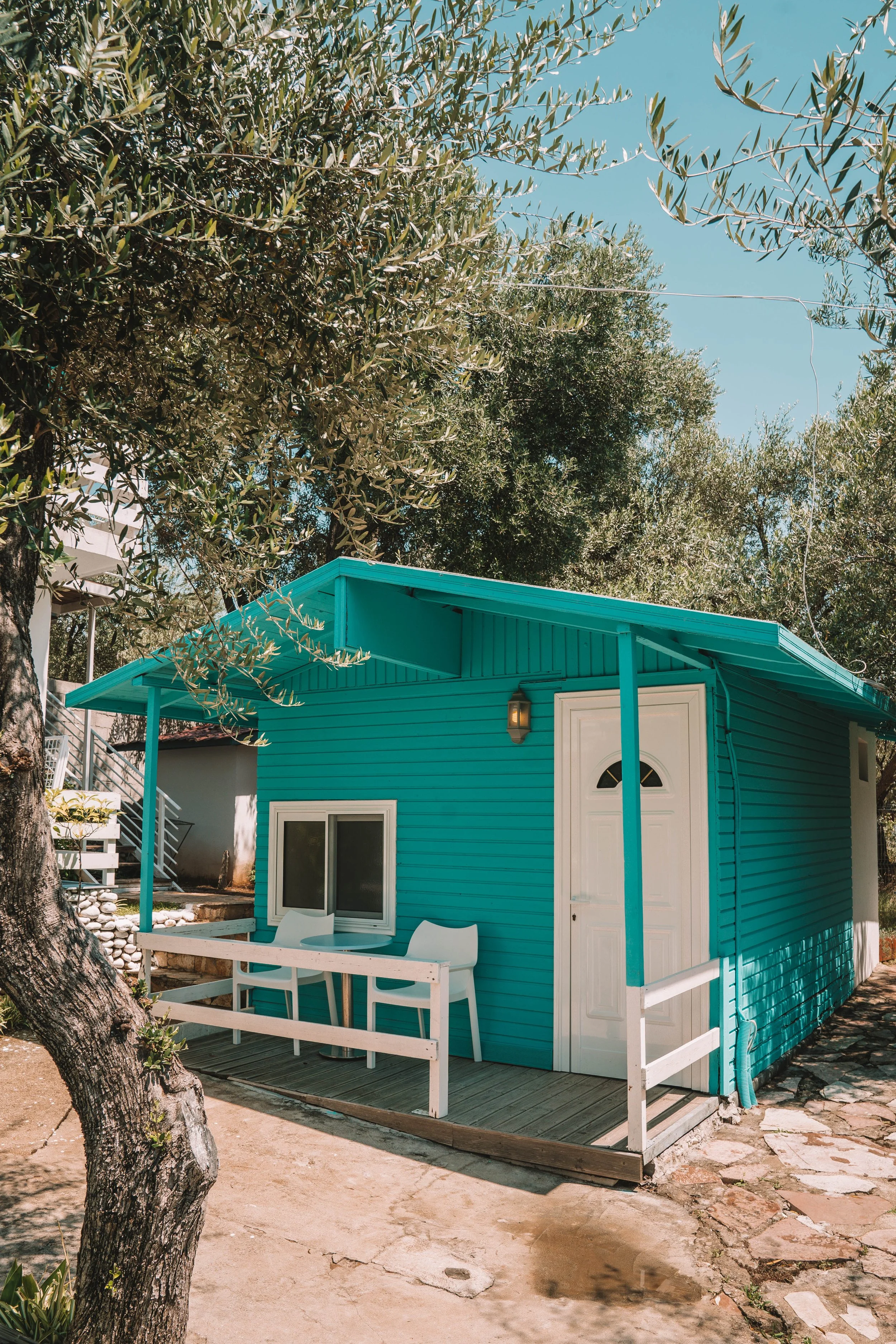 Small turquoise cabin with white door and railing, surrounded by trees and a stone path.