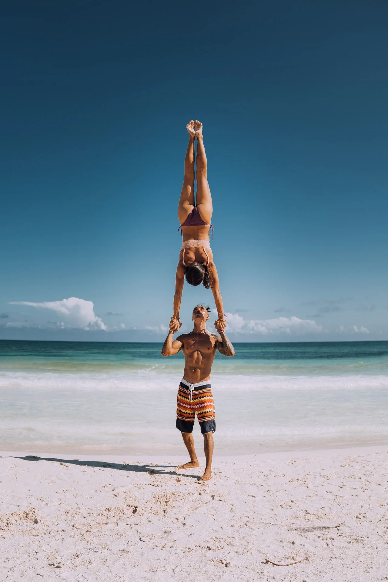 Two people performing an acrobatic handstand on a beach, with clear blue sky and ocean in the background.