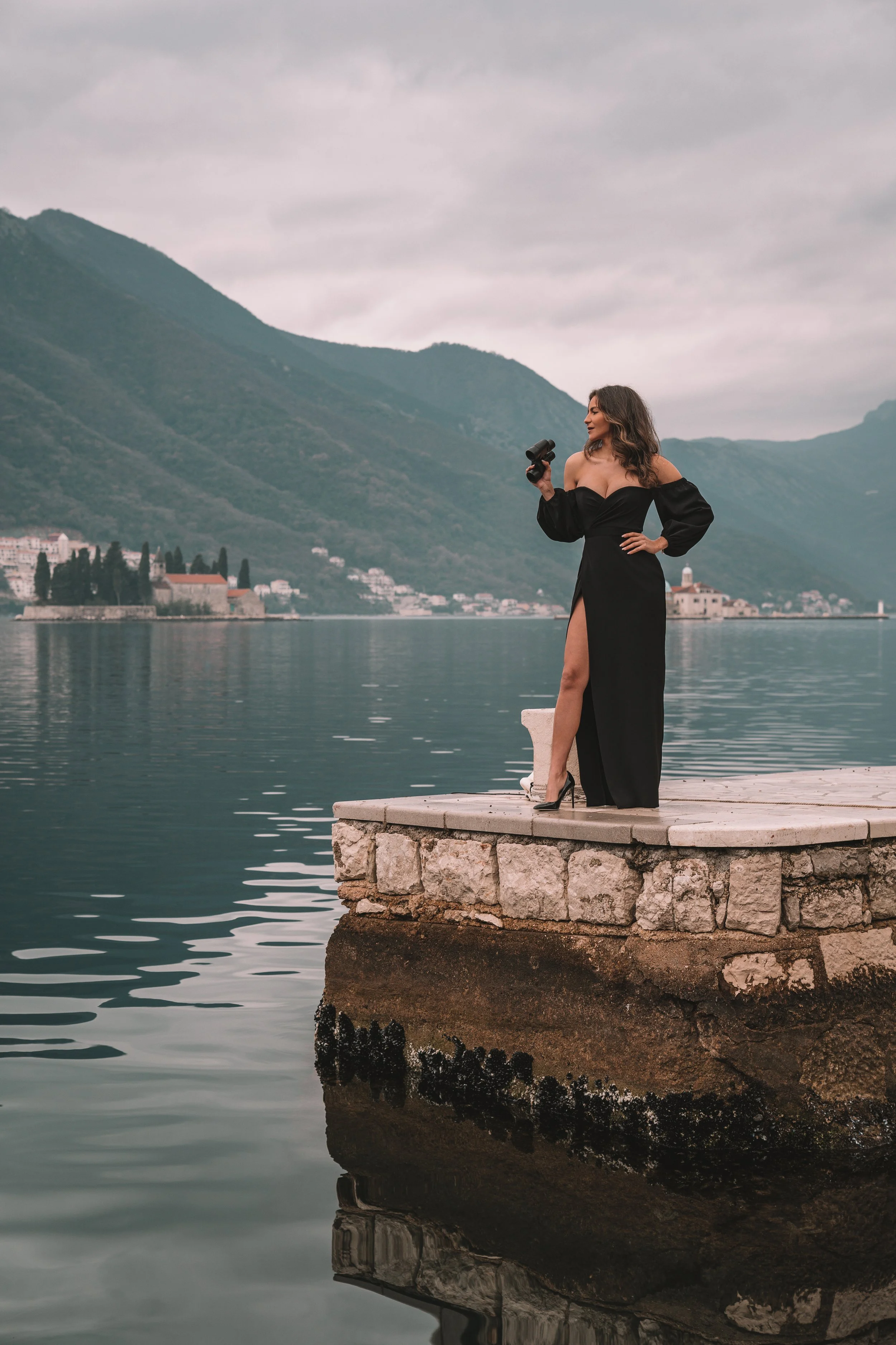 Woman in black dress holding a camera standing on a stone pier by a calm lake with mountains and buildings in the background.