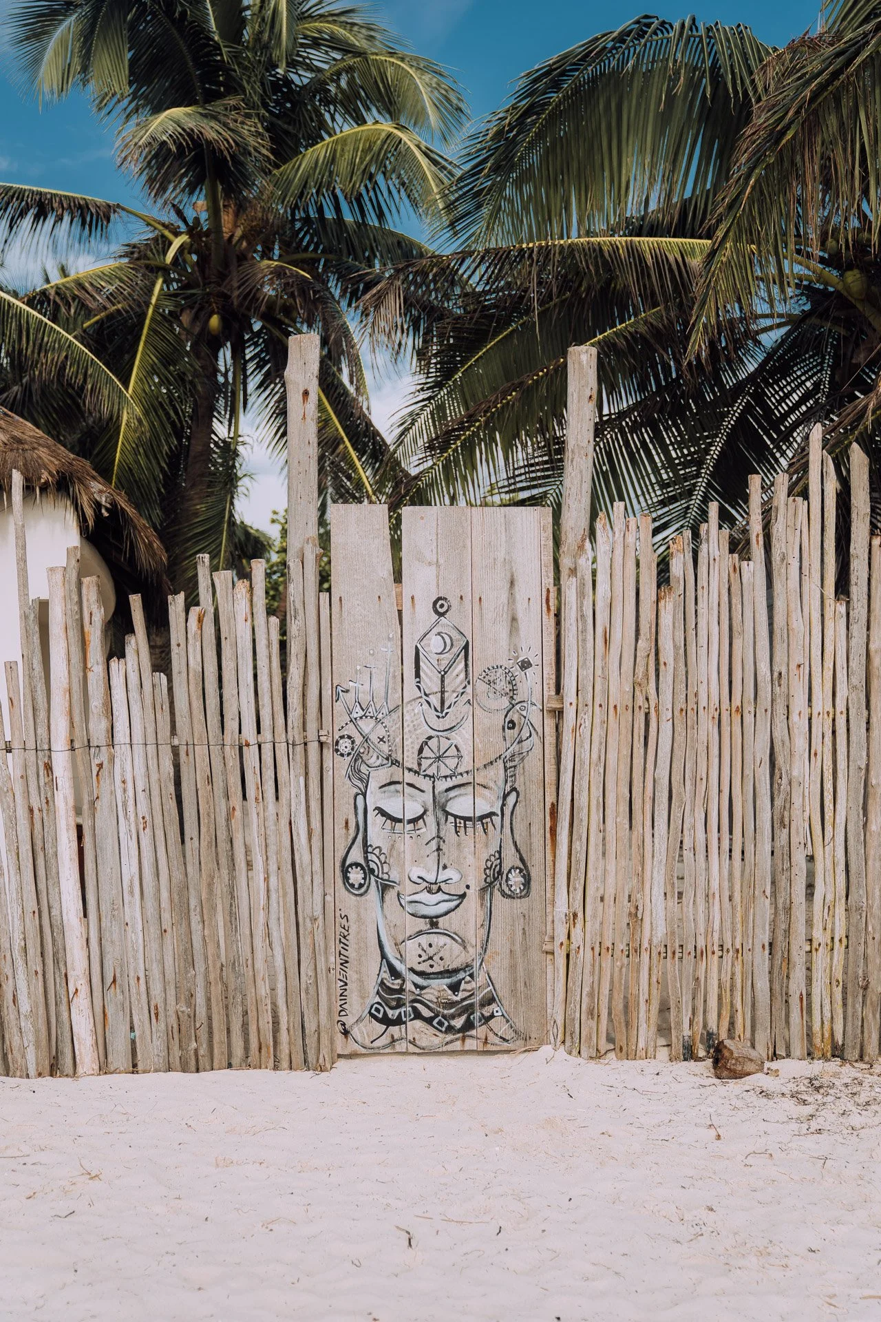 Wooden fence with tribal art in front of palm trees on a beach.