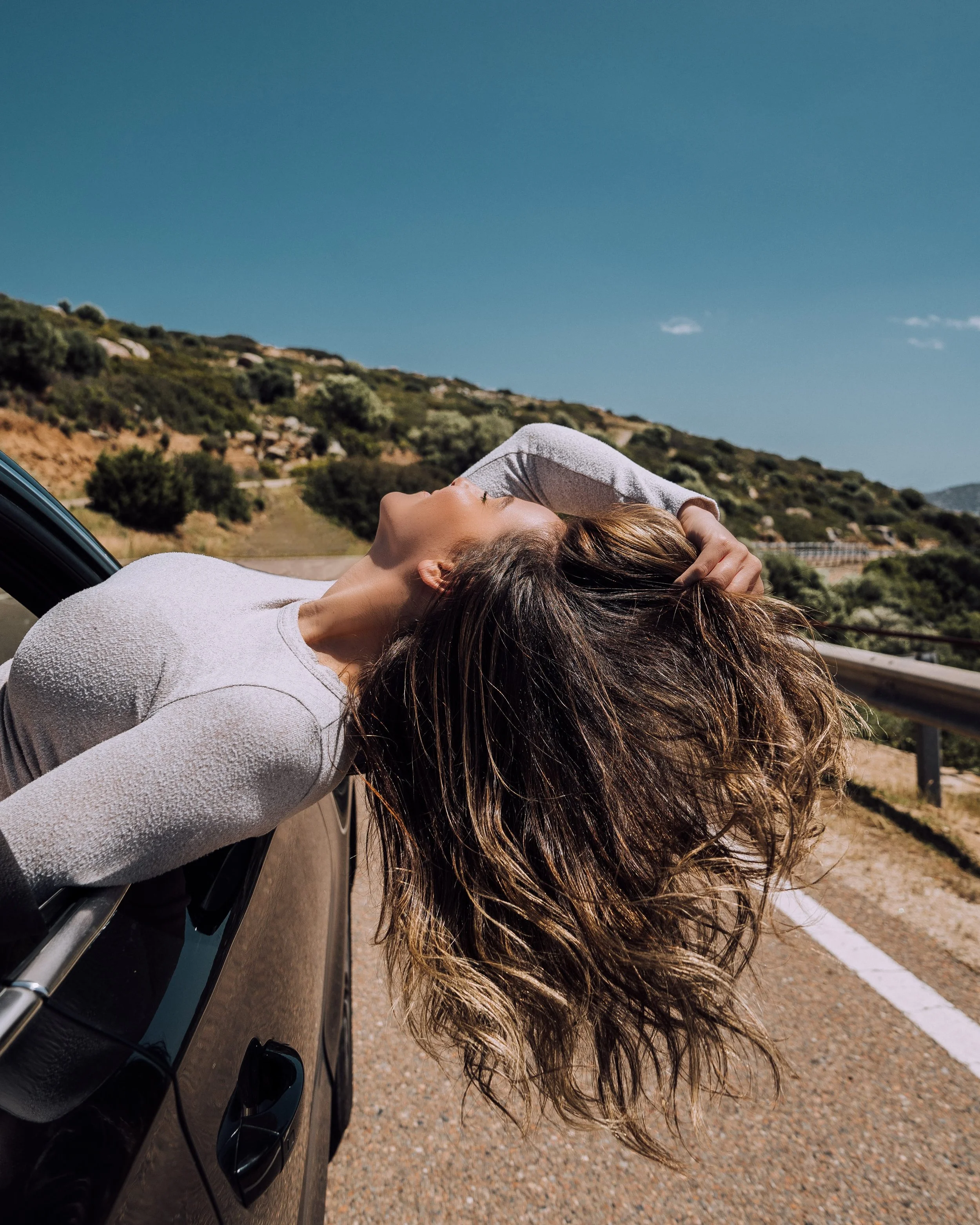 Person leaning out of a car window with head tilted back, long hair flowing in the wind, driving on a road through a scenic landscape with hills and bushes, under a clear blue sky.
