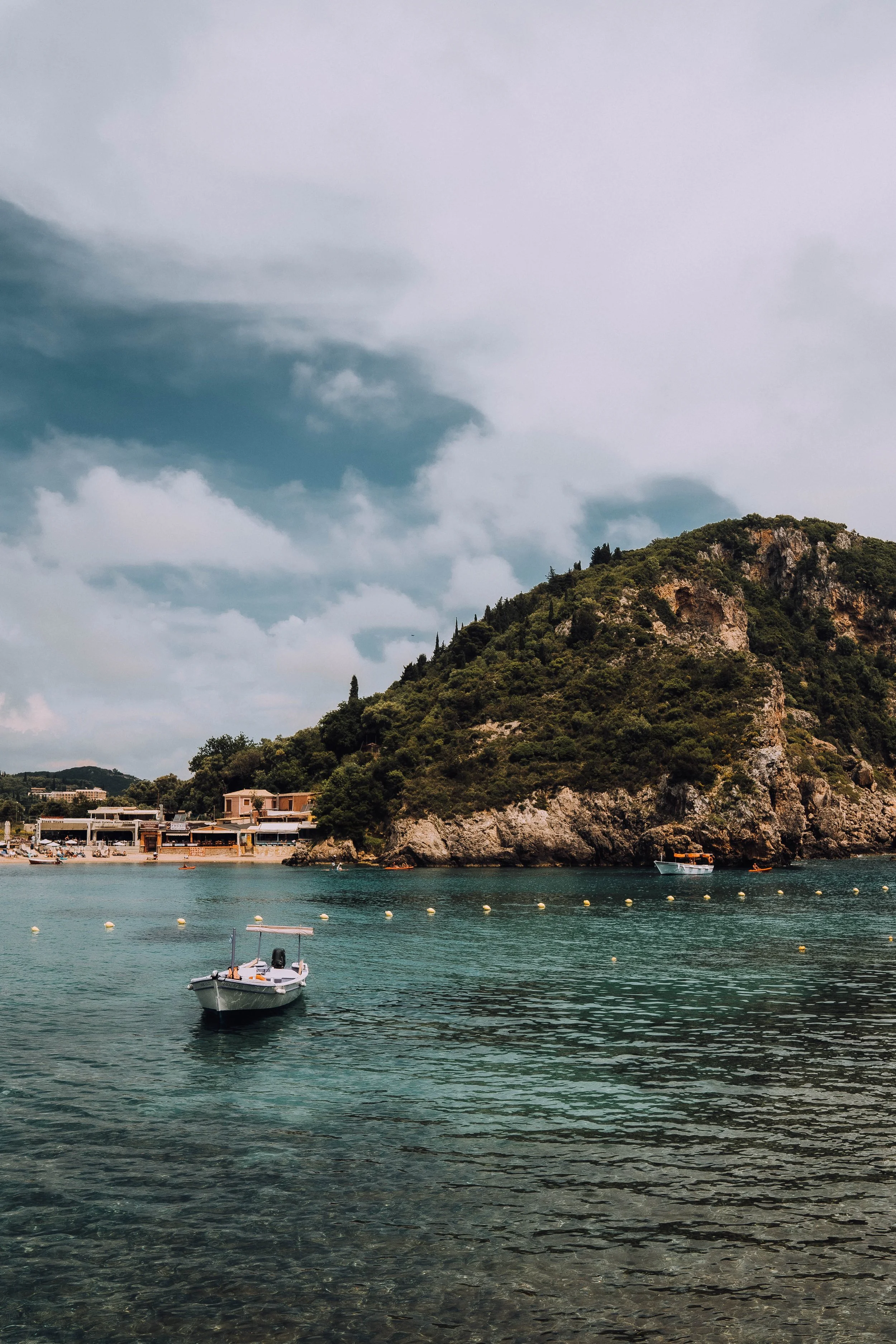 Scenic coastal view with a small boat on turquoise water, surrounded by rocky cliffs and a hill covered in greenery under a cloudy sky.