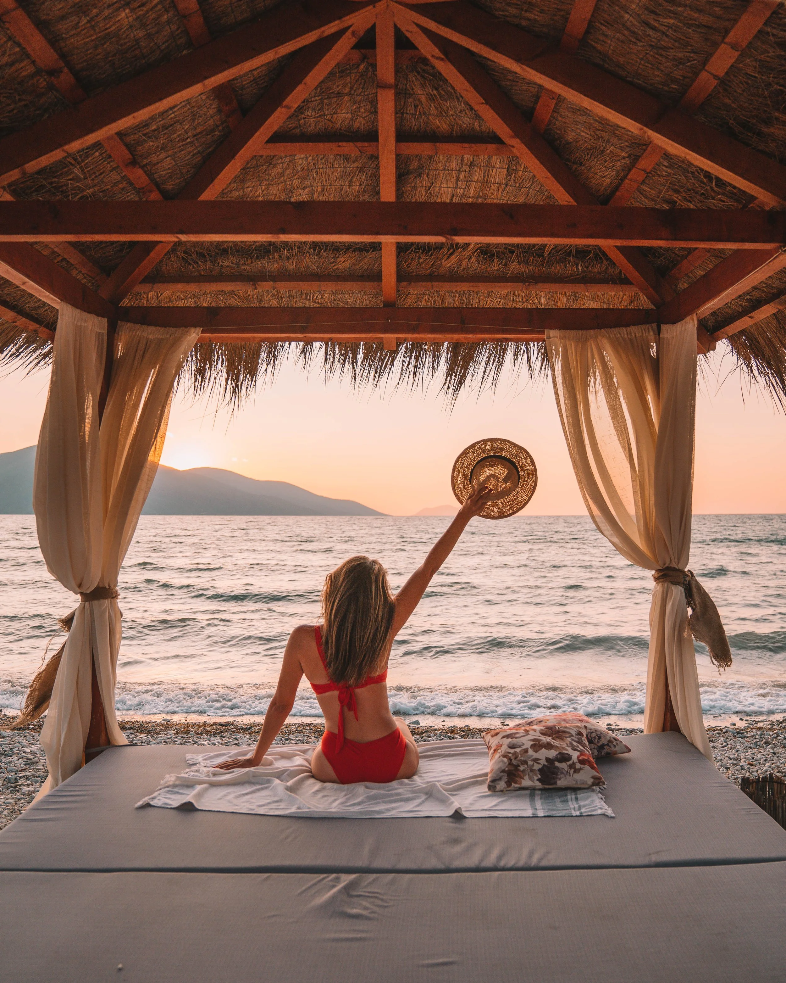 Woman in red bikini sitting under gazebo on beach at sunset, holding hat, facing ocean.