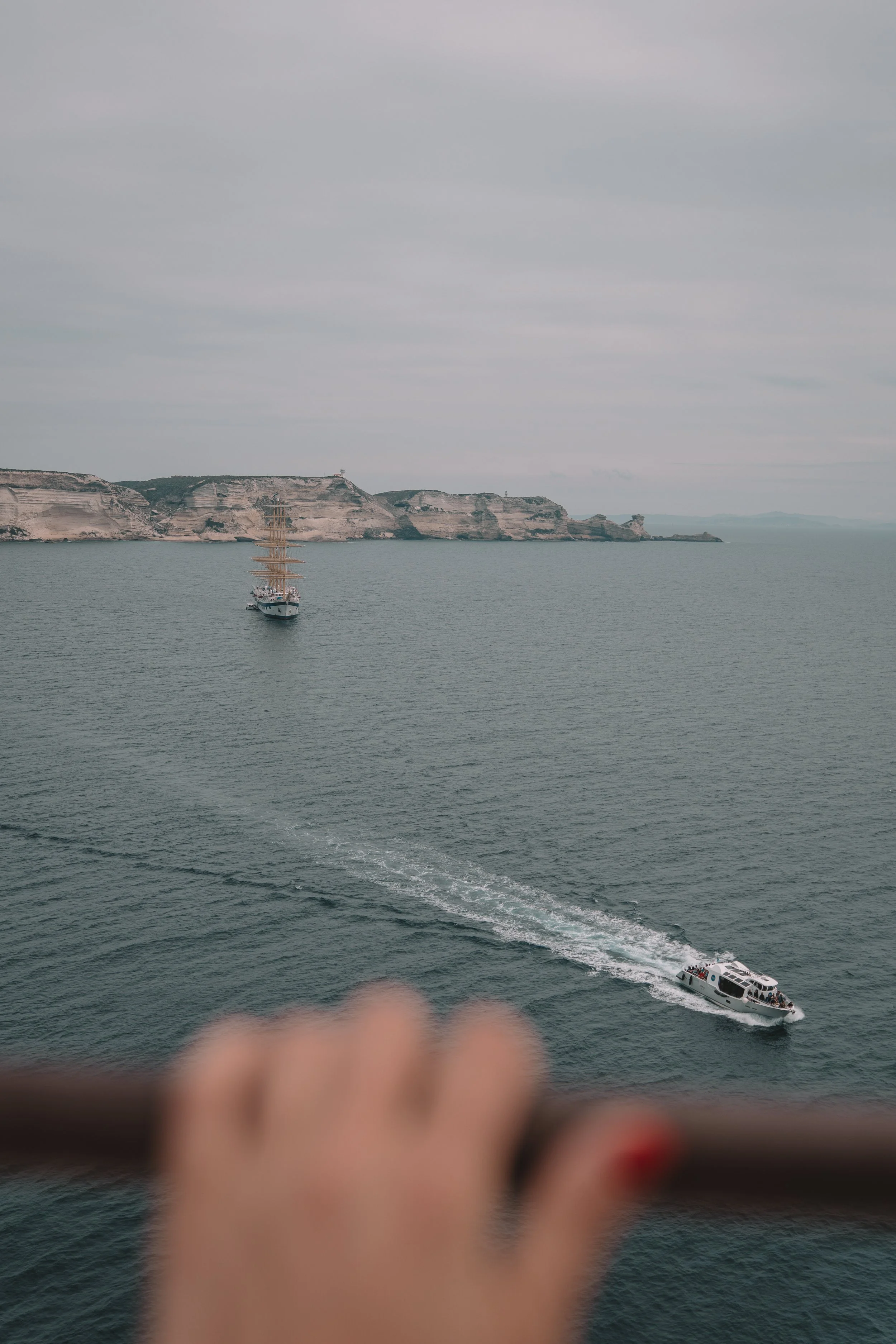View of a seascape with two boats on the water, a large sailboat and a smaller motorboat, with cliffs in the background and a blurred hand on a railing in the foreground.