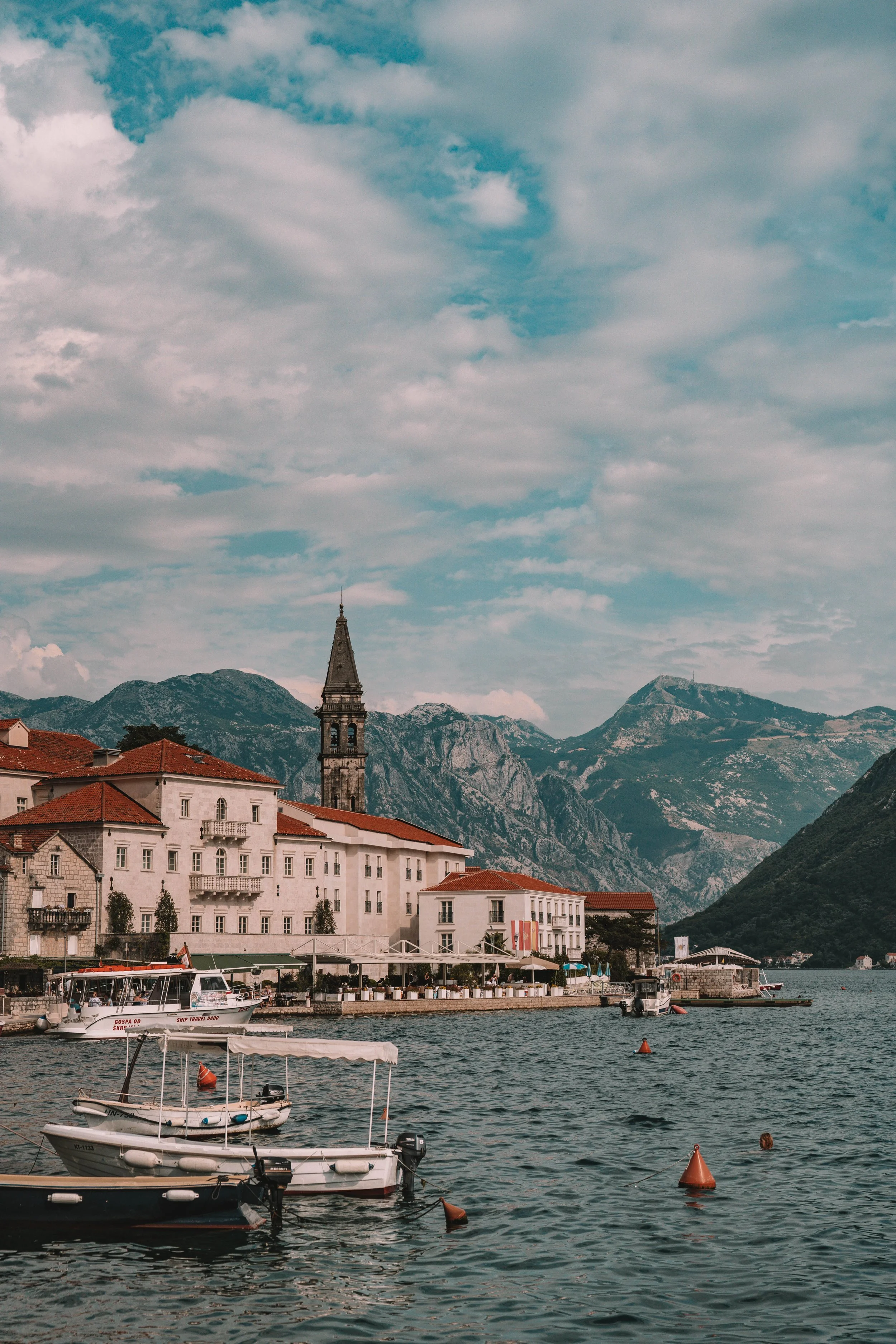 A scenic view of a coastal town with red-roofed buildings and a bell tower by the water, surrounded by mountains under a partly cloudy sky.