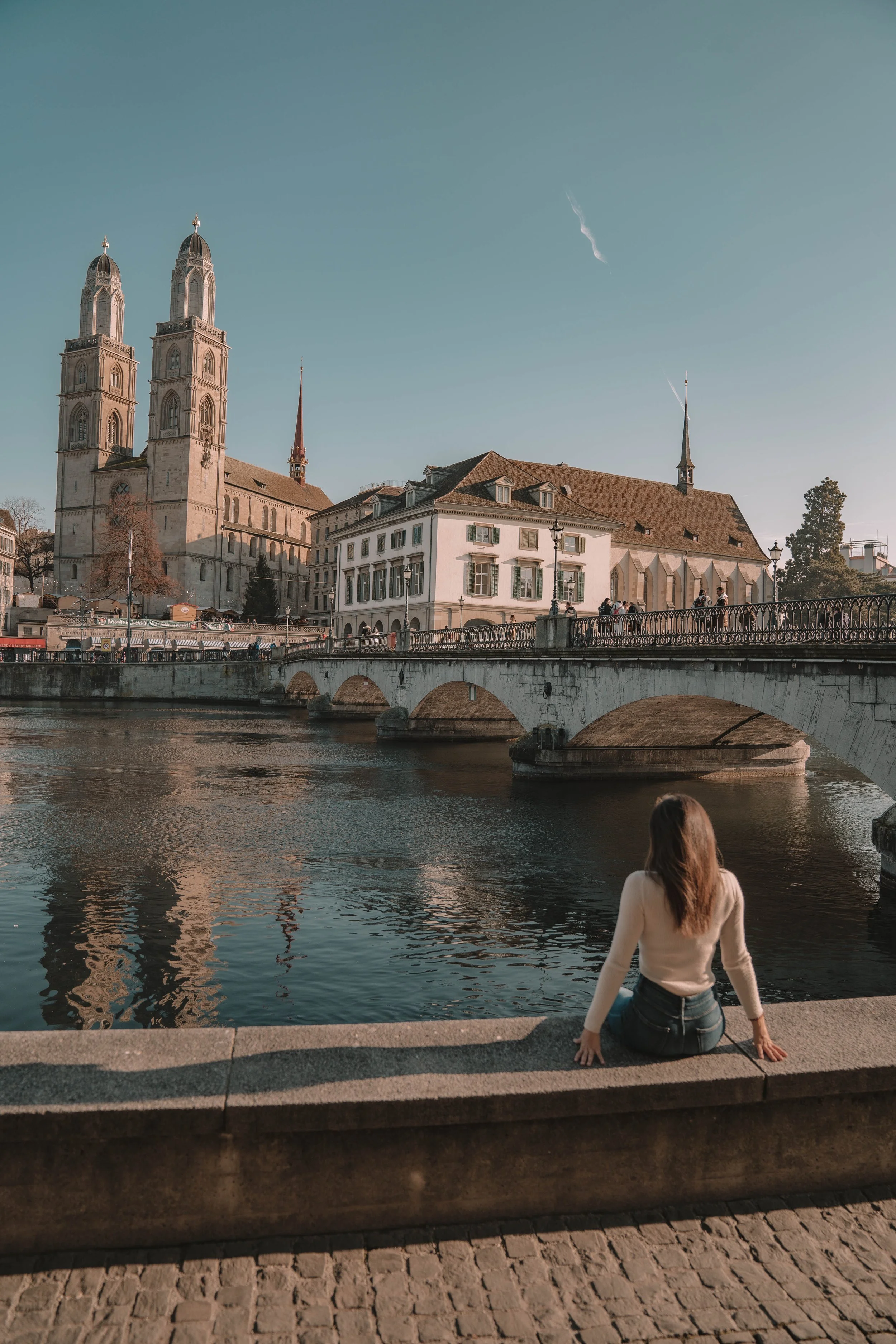 Woman sitting by river with historic buildings and bridge in the background.