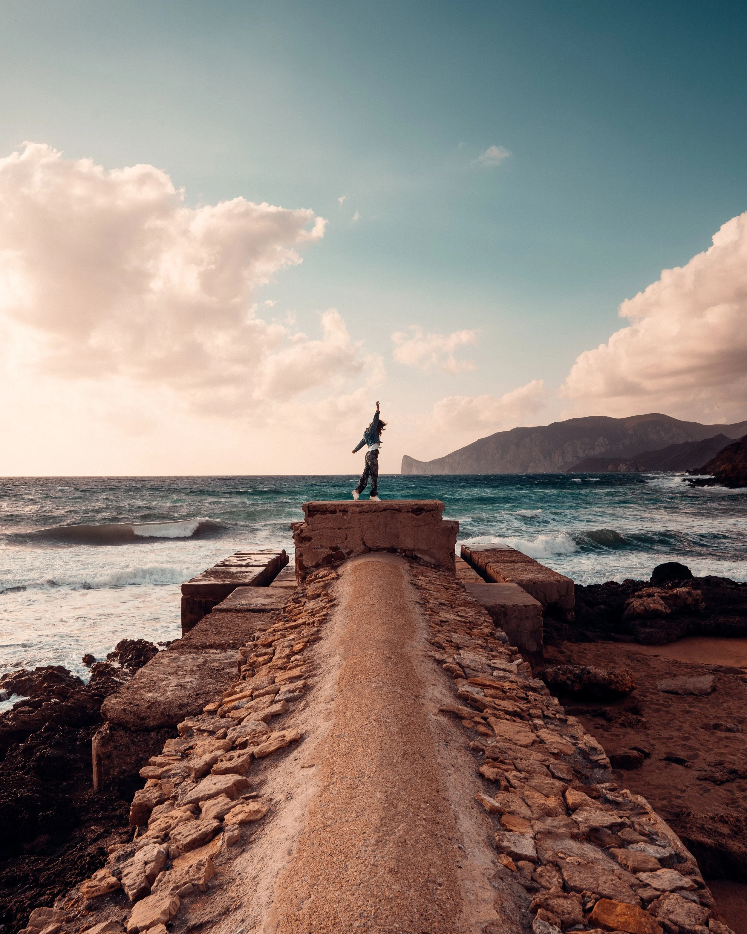 Person standing on a rocky pier overlooking the ocean, with hills in the background and a partly cloudy sky above.