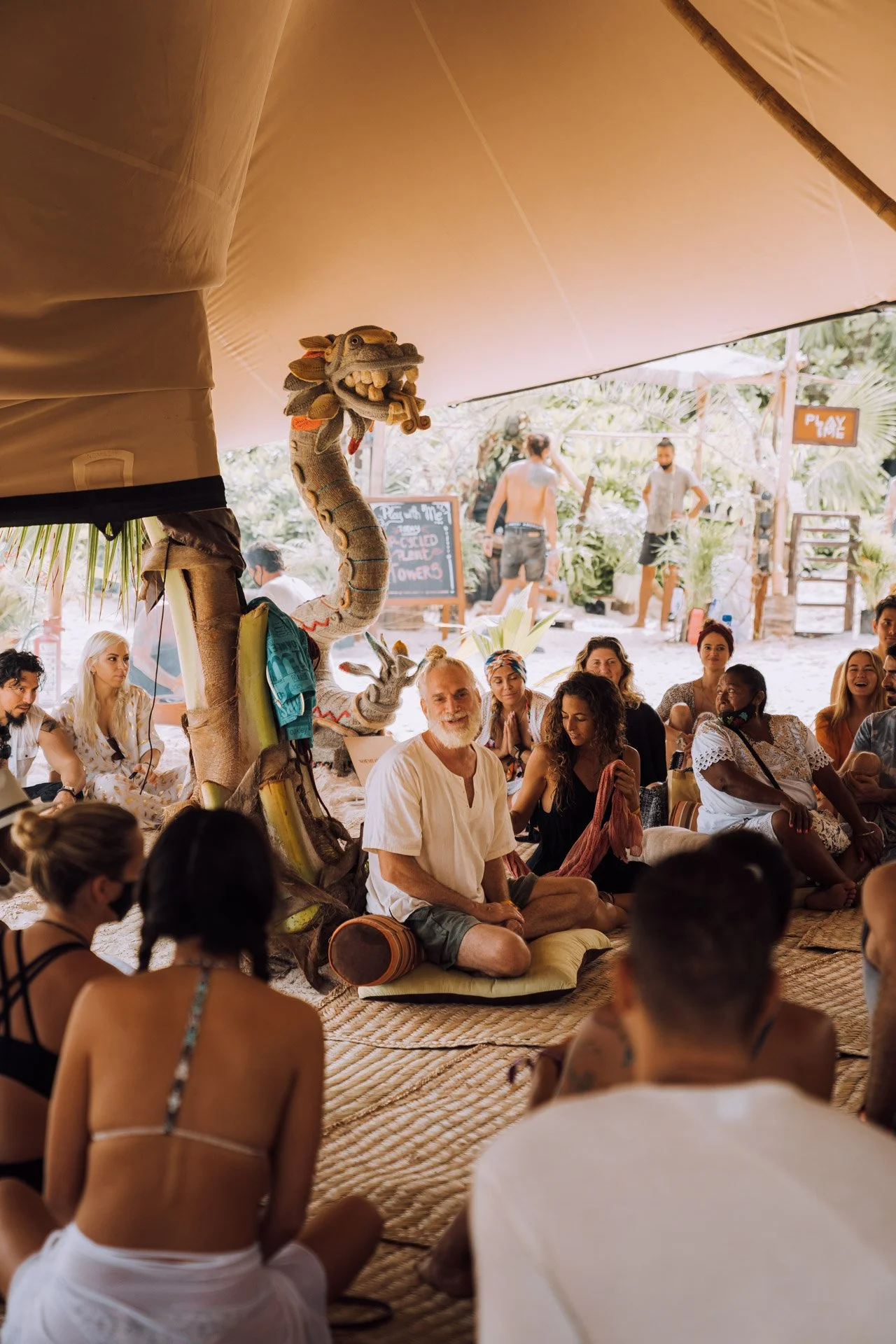 A group of people sitting on mats under a tent, attending a gathering or workshop. A wooden dragon sculpture is present. The setting appears relaxed and informal, with a mix of casual clothing and tropical surroundings.