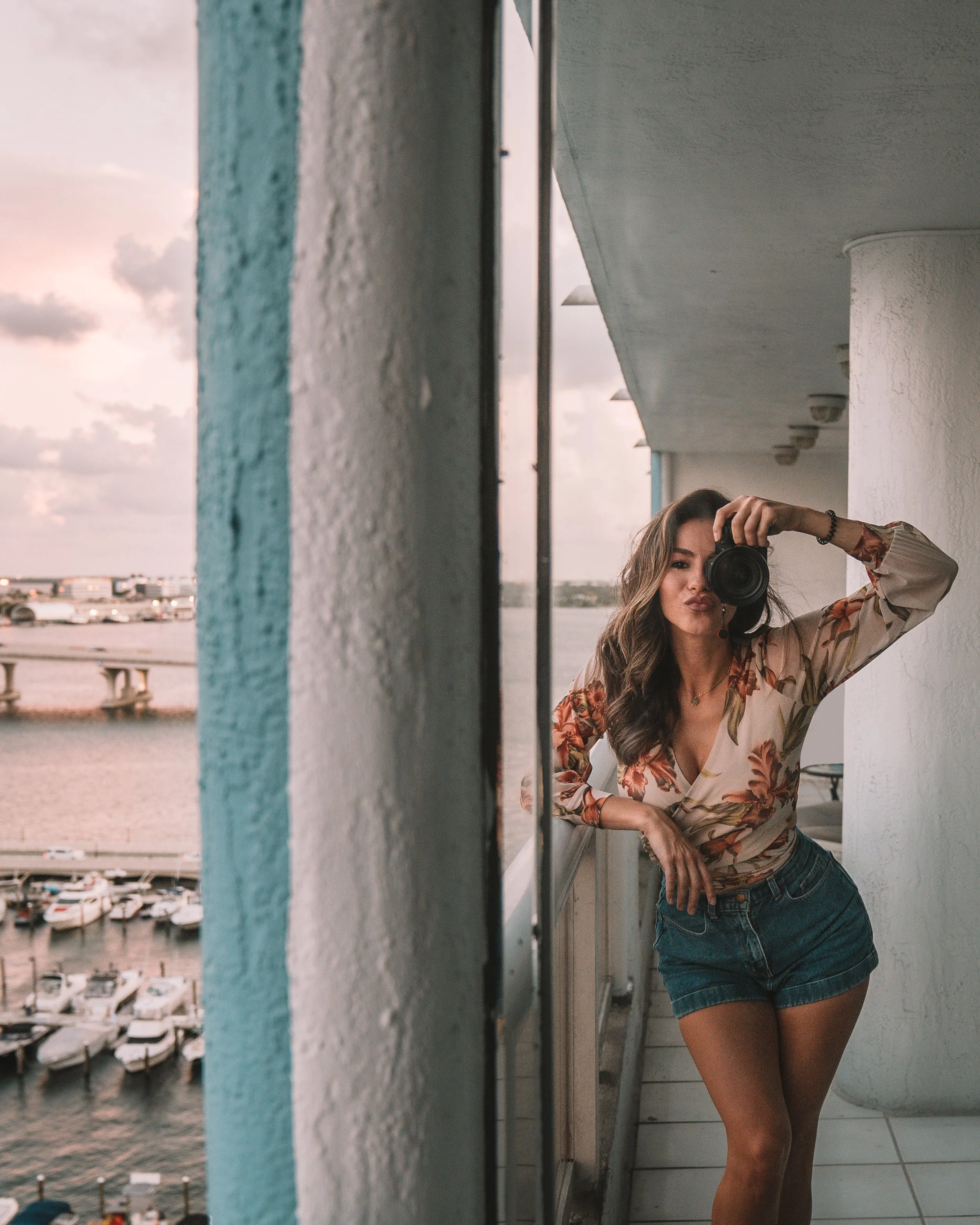 Woman taking a selfie in a mirror on a balcony overlooking a marina with boats. She is wearing a floral top and denim shorts, holding a camera to her face.