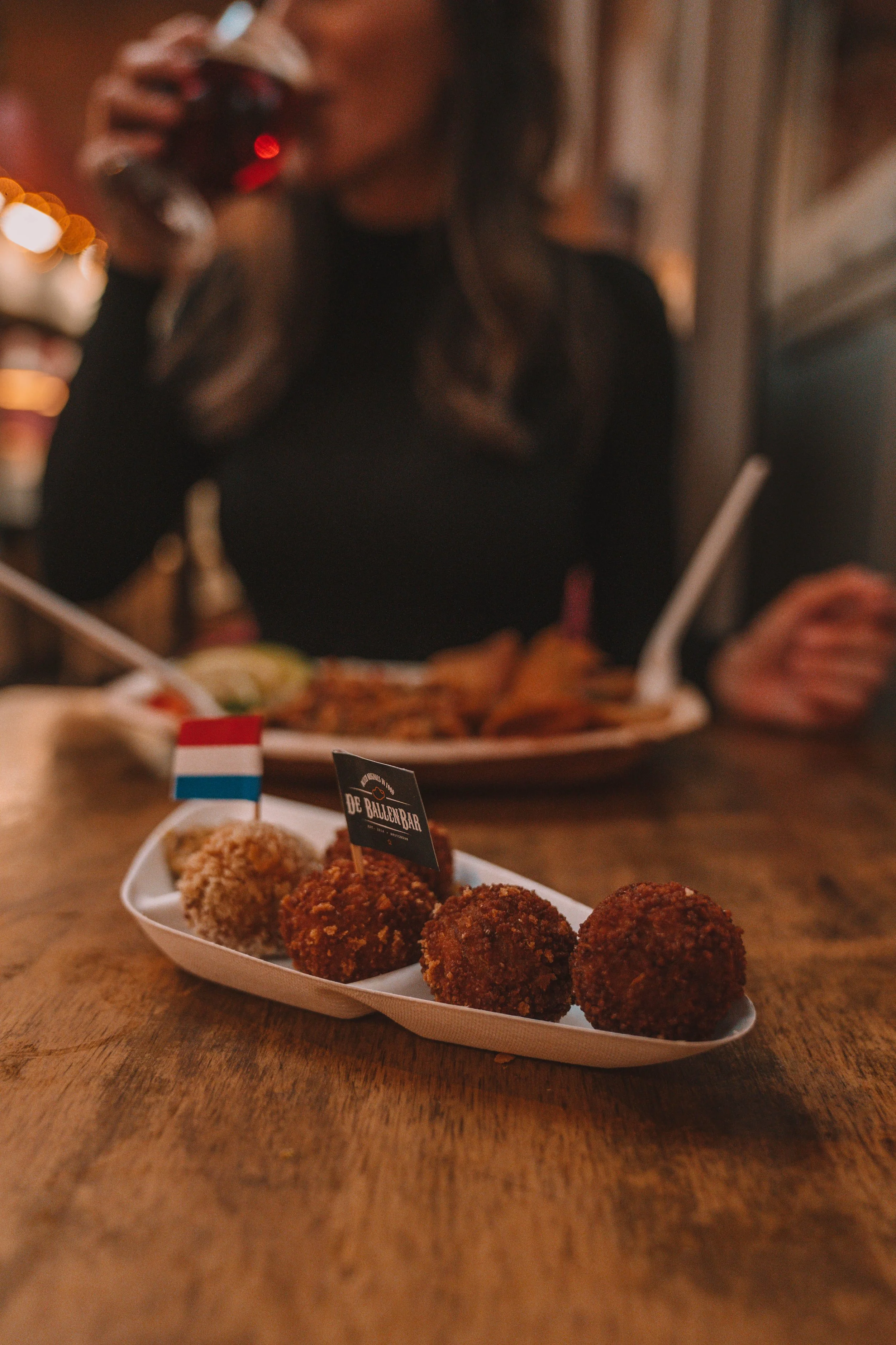 Person in black outfit sipping a drink with a plate of various fried food balls on the table, accompanied by a small Dutch flag and a label saying "De Ballen Bar."