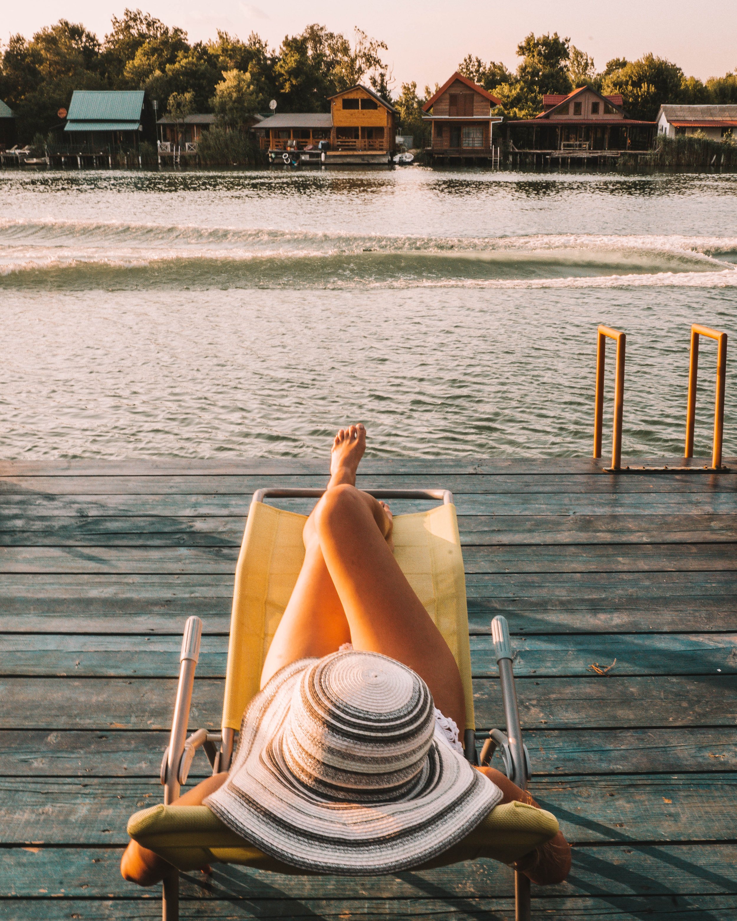 Person relaxing in a lounge chair on a wooden dock by a lake, wearing a wide-brimmed hat. In the background are trees and lakefront cabins.