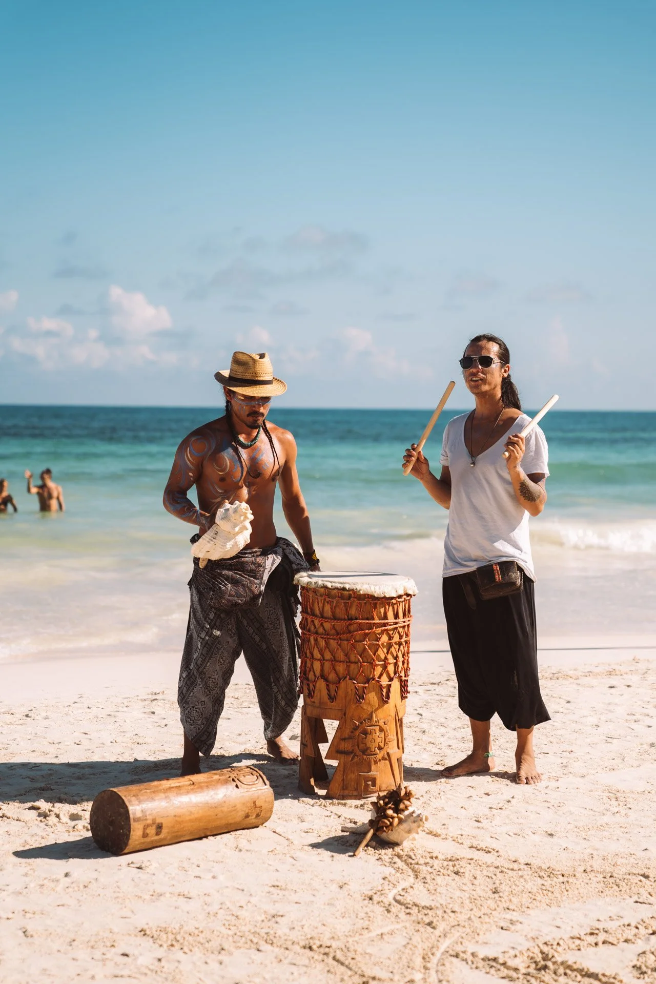 Two men playing traditional drums on a sandy beach with the ocean in the background, one man wearing a straw hat and patterned pants, the other in a white shirt and holding drumsticks. Tulum, Mexico