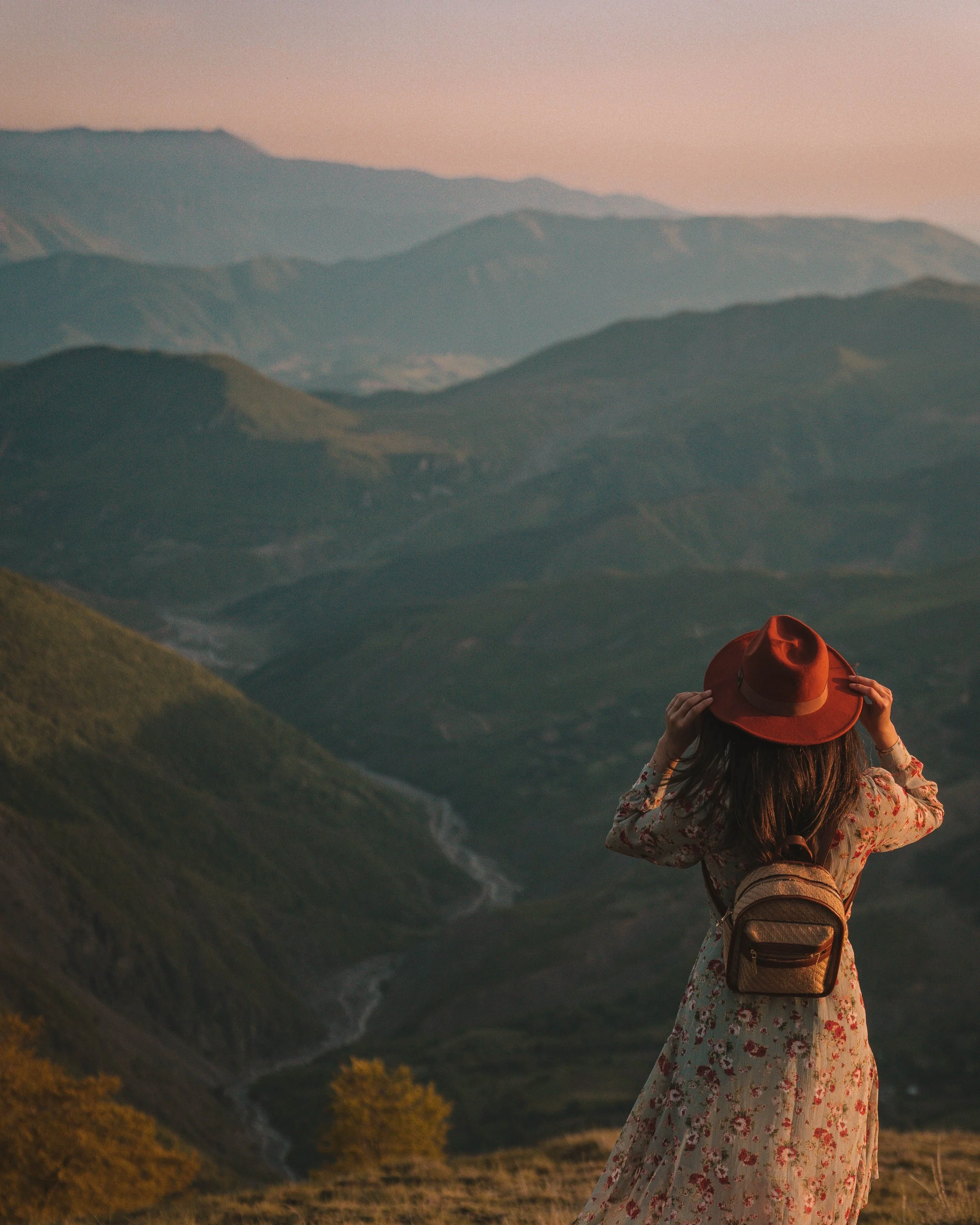 Woman in floral dress and red hat overlooking mountains during sunset.