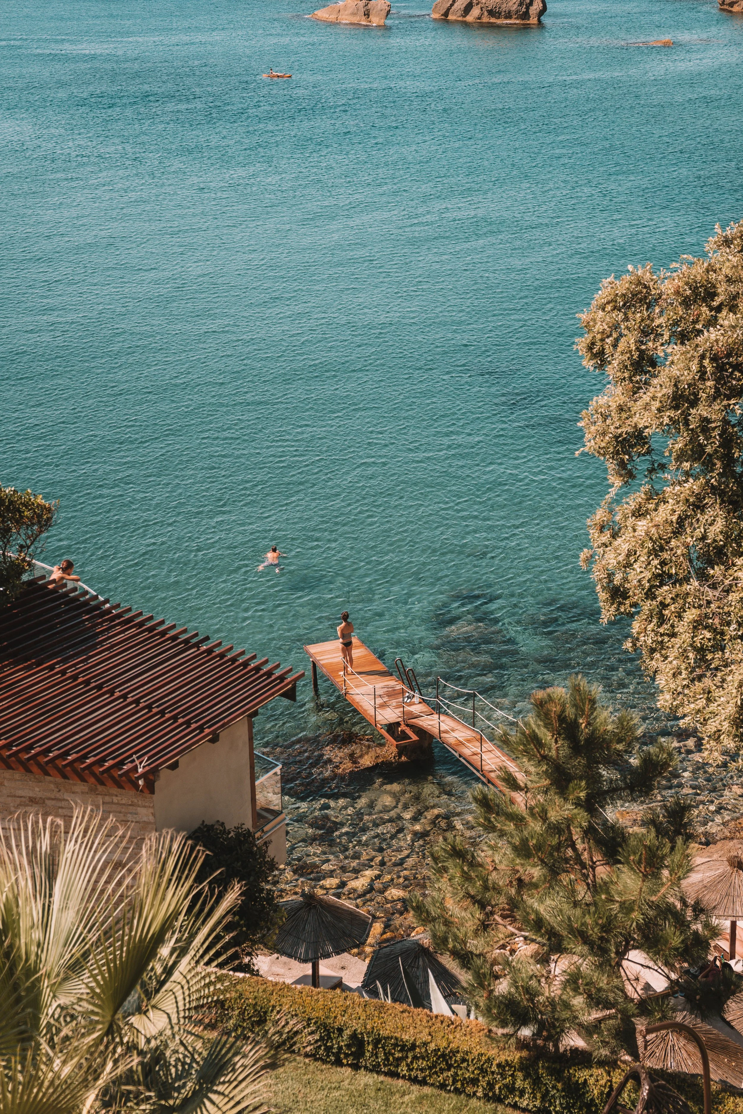Scenic coastal view with clear blue water, a wooden pier, swimmers, lush greenery, and rocky outcrops in the distance.