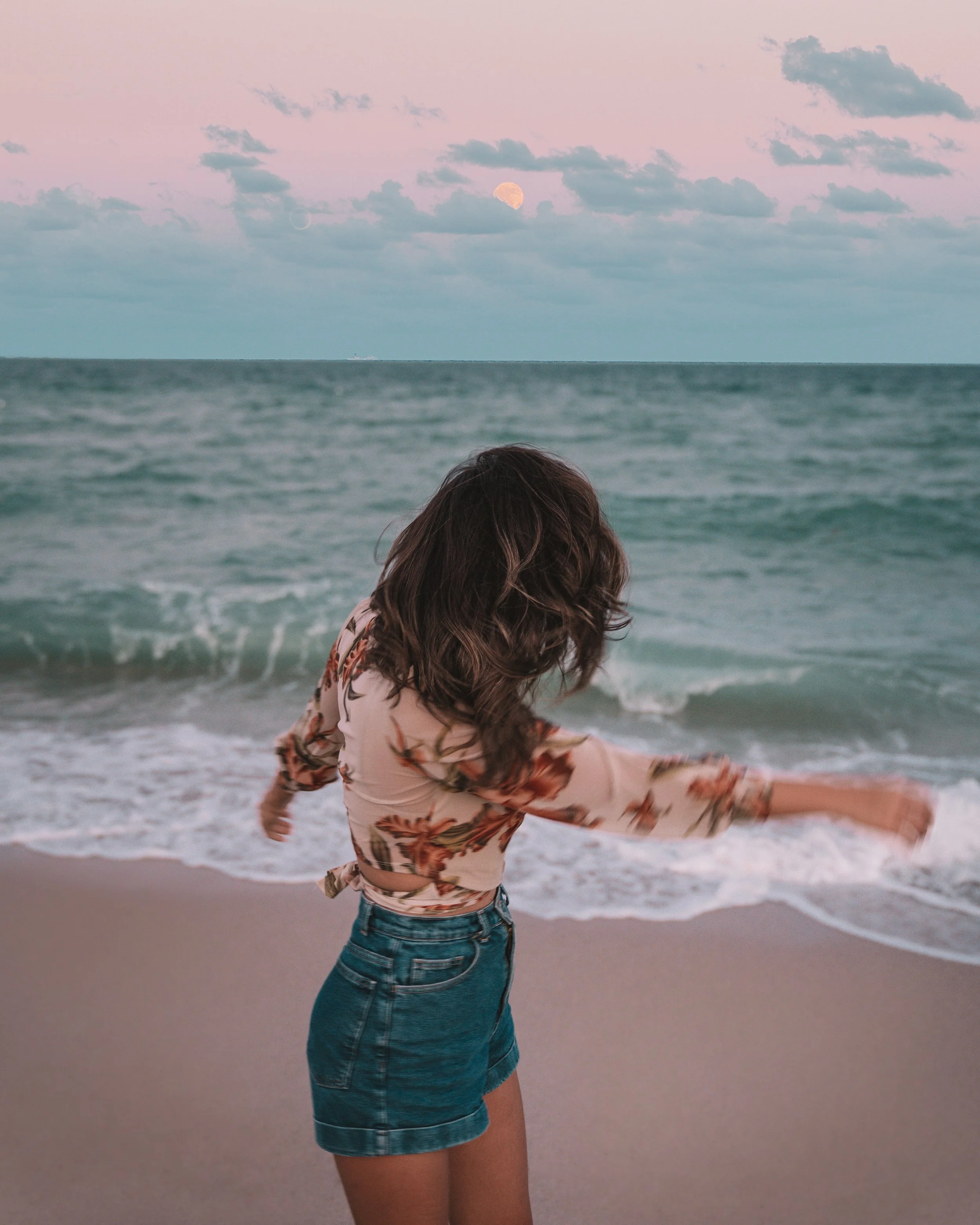Woman on beach in floral top and denim shorts, with ocean waves and sunset in the background.