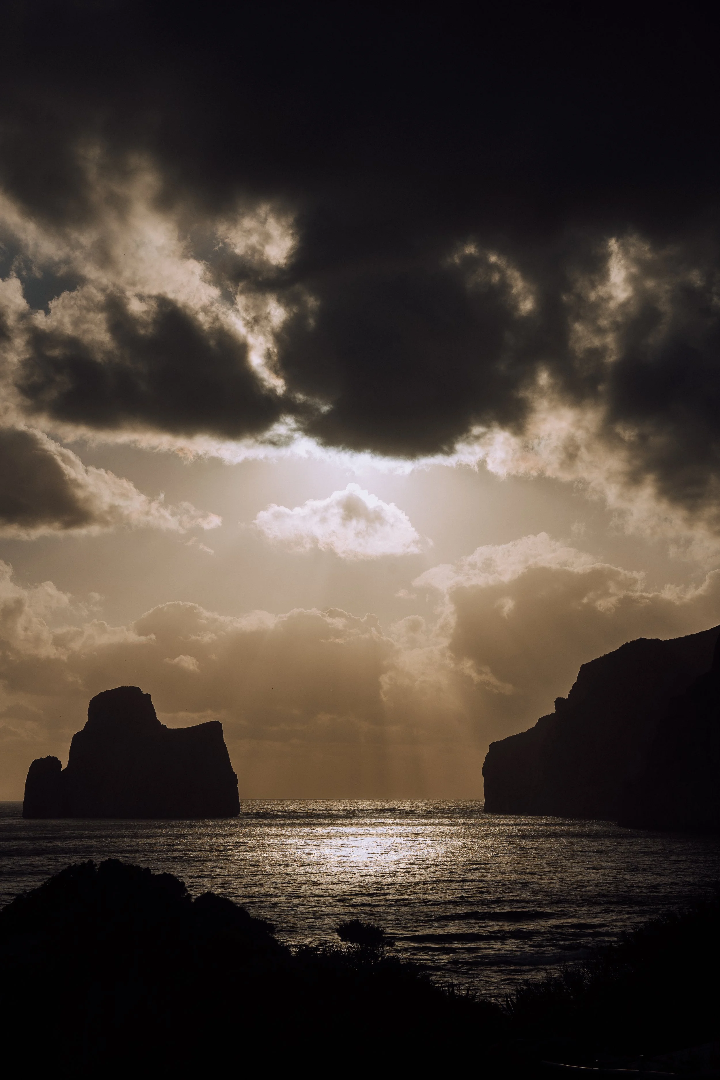 Silhouette of a rocky coastline at sunset with dramatic clouds and shimmering ocean water.