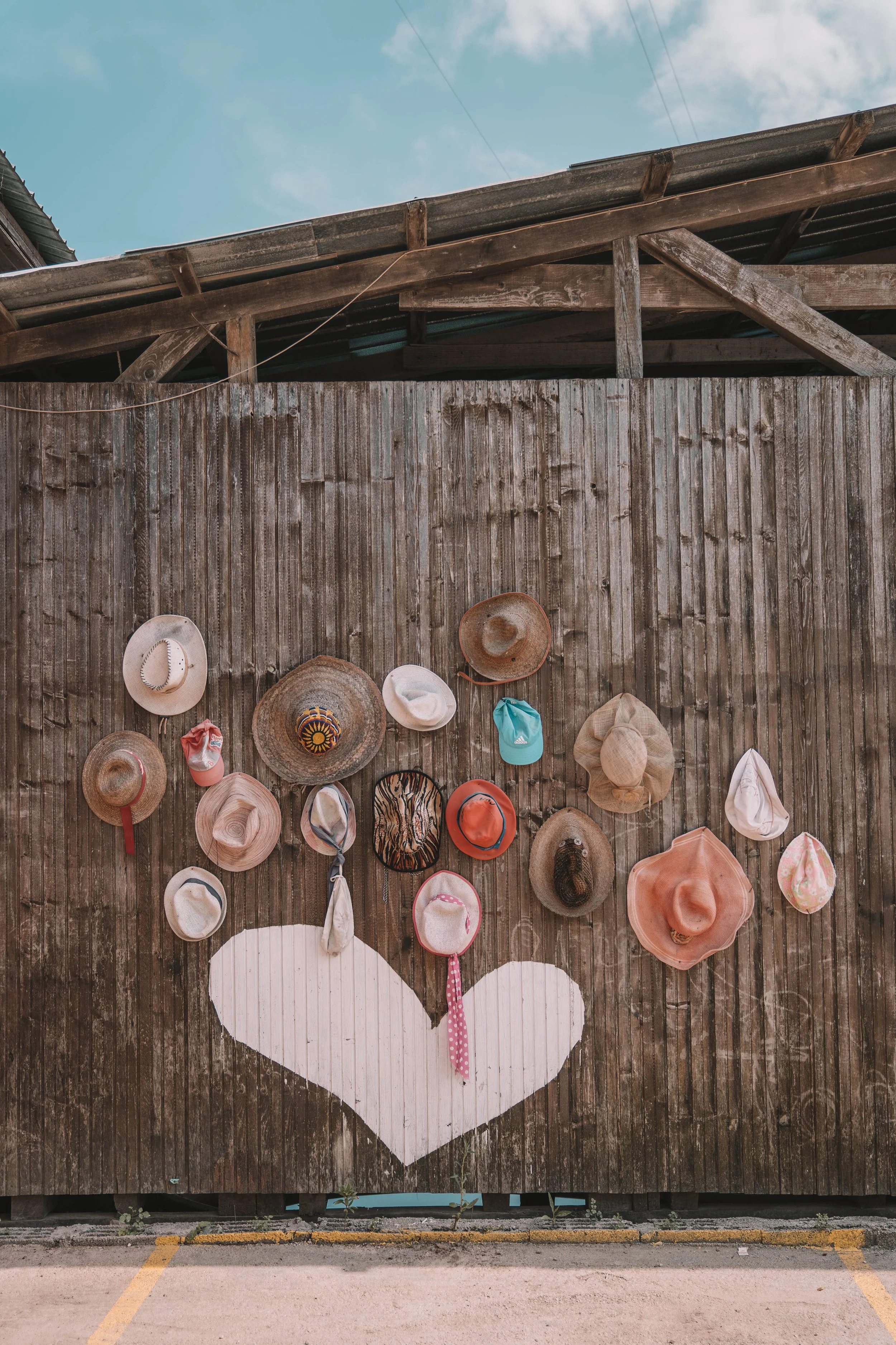 A rustic wooden wall displays various hats, including straw and fabric types, arranged in a loose heart shape around a painted white heart on the planks.