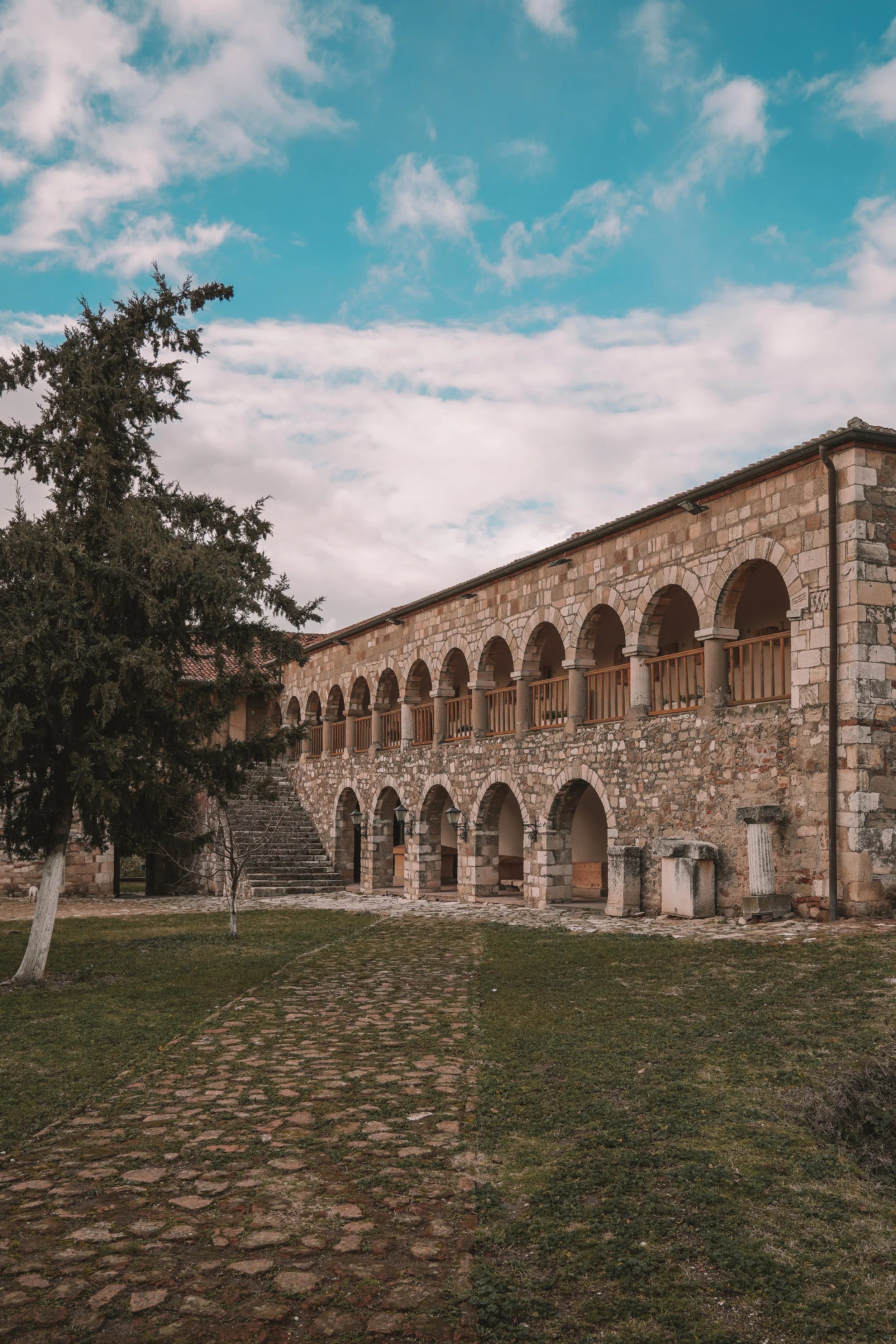 Stone building with arched openings and an outdoor staircase, surrounded by grass and a tree, set against a partly cloudy sky.
