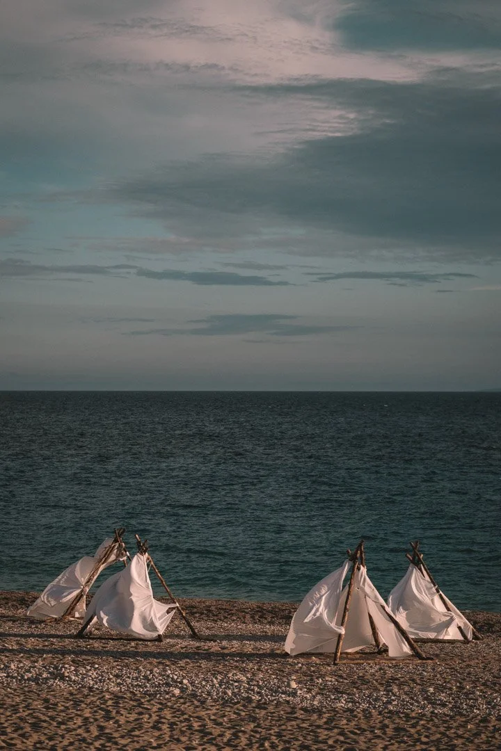 Beach with teepee-like tents, ocean in background, cloudy sky.