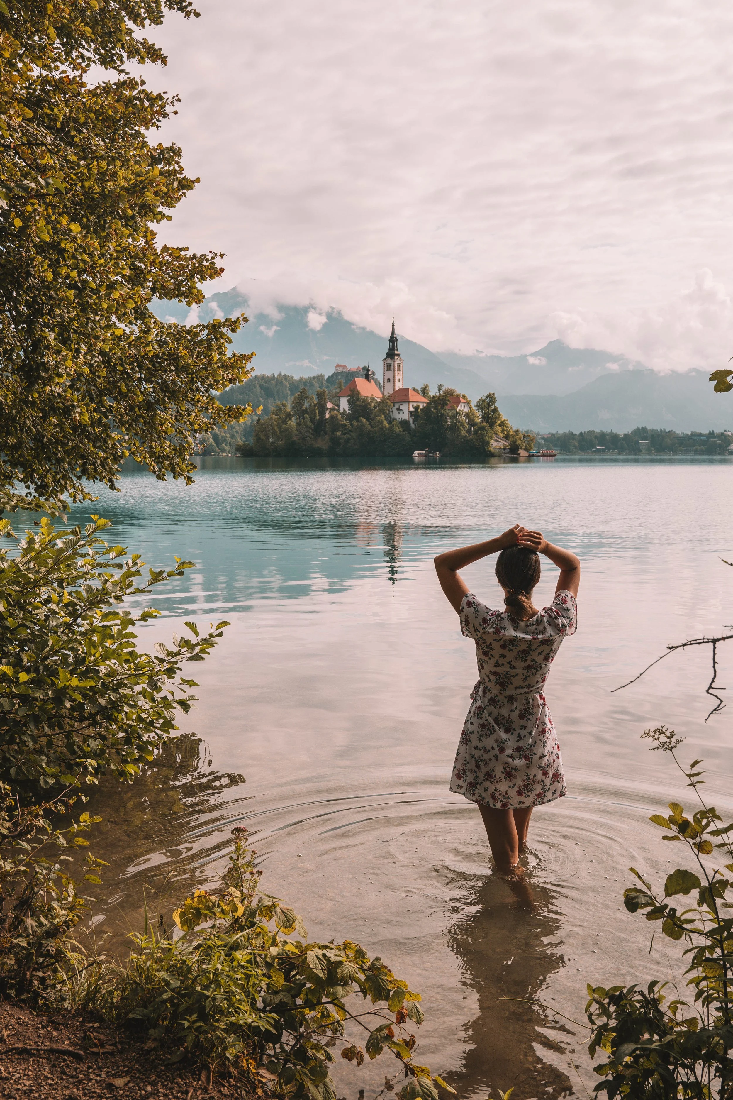 Person standing in a lake, facing a church on an island, with trees and mountains in the background.