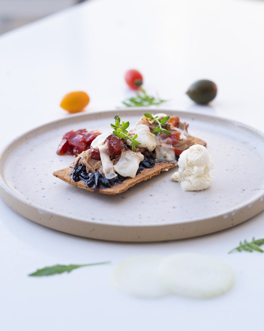 Gourmet appetizer featuring artisan cracker, melted cheese, cured meat, and fresh herbs, styled on a ceramic plate with cherry tomatoes and cauliflower — photographed in Albania by Albanian female photographer based in New York.